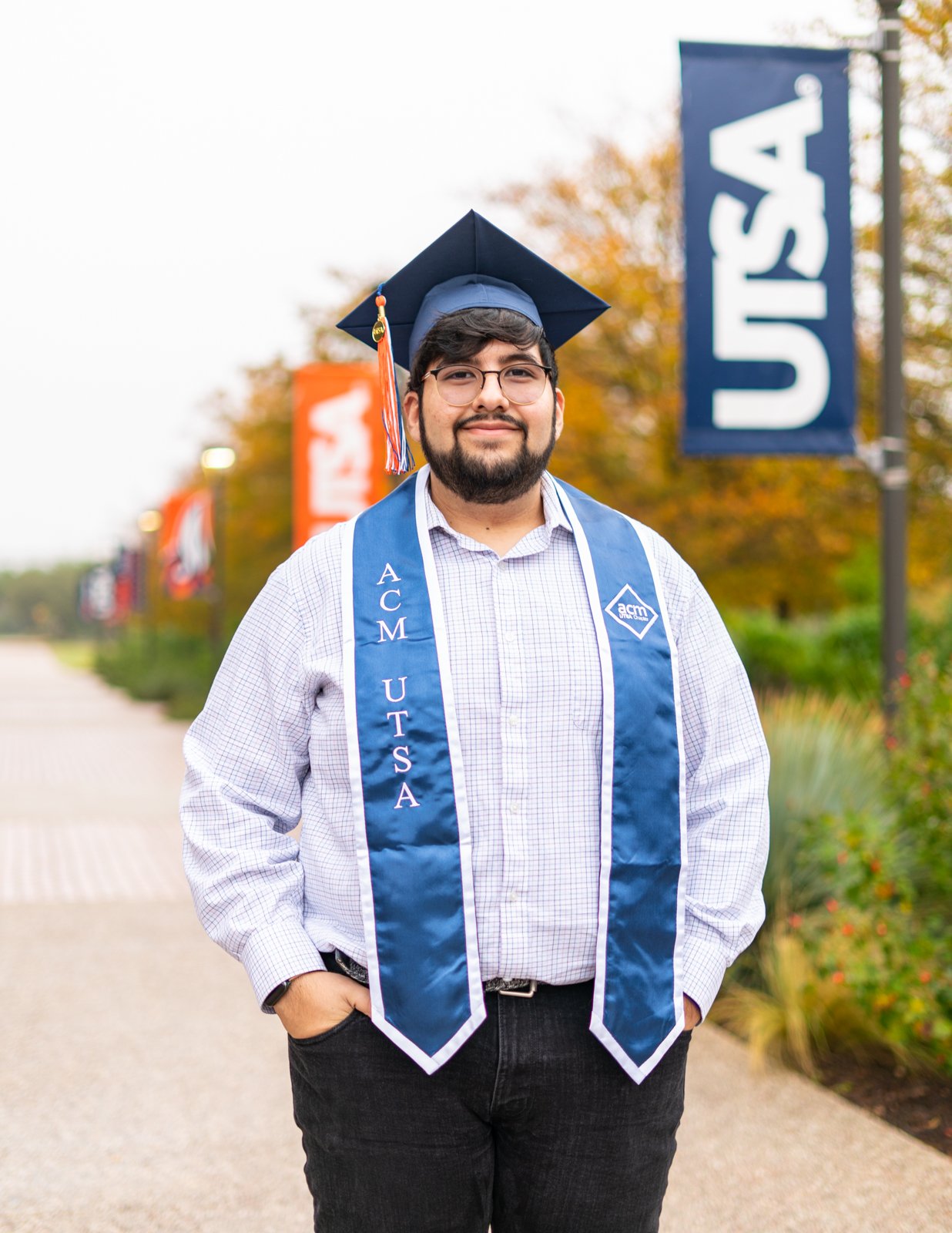 A young man wearing glasses, a graduation cap, and a blue sash that reads 'ACM OUTSA,' standing outdoors in front of a blue 'USA' banner with autumn trees in the background.