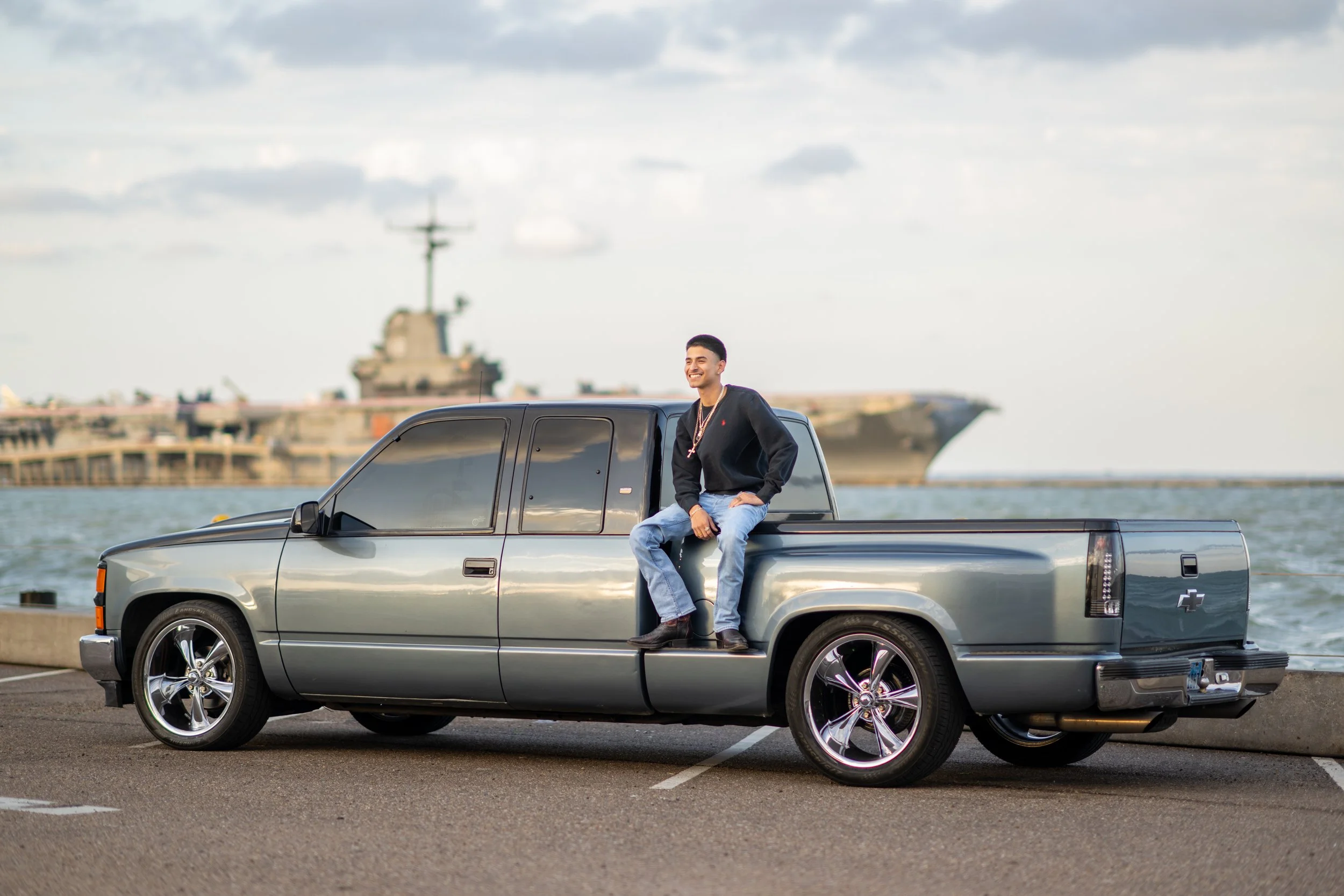 A smiling young man in a black jacket and jeans sitting on the edge of a silver Chevrolet pickup truck, parked near a waterfront with a large naval ship in the background on a cloudy day.