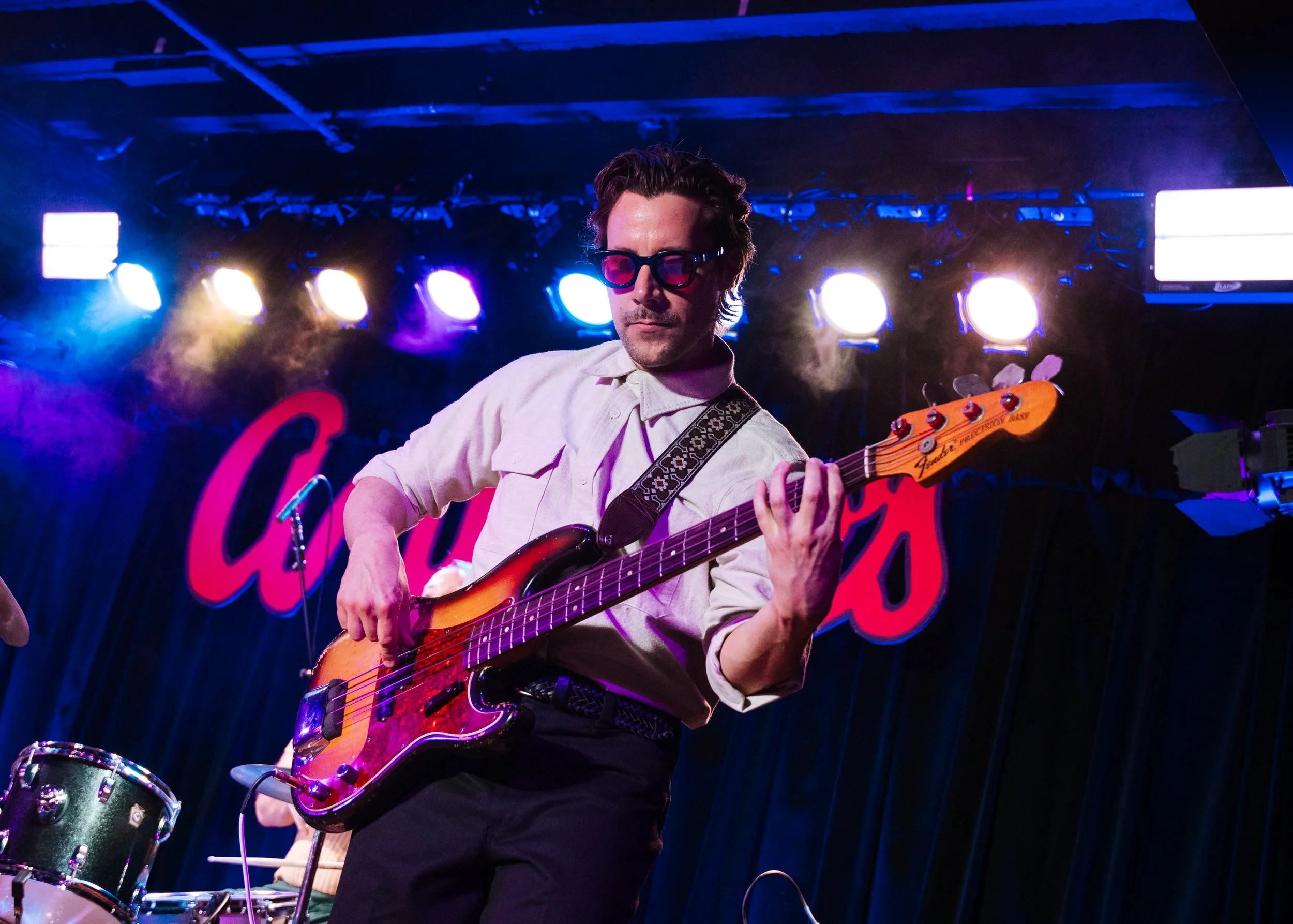 A man playing an electric bass guitar on stage with colored stage lights and a dark background.