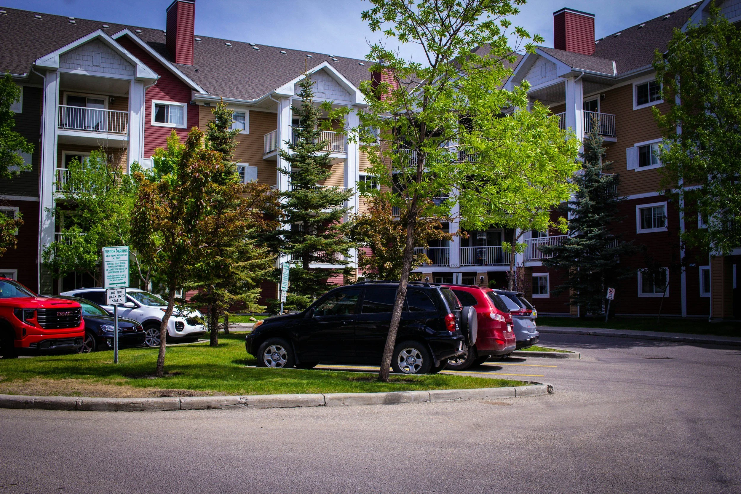 A parking lot in front of a multi-story residential building with trees and parked cars, and parking signs visible.