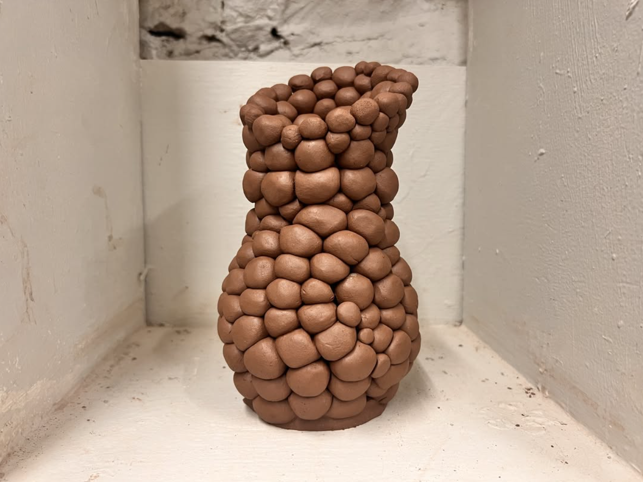 A tall vase made of small brown clay balls, placed on a white surface in a corner with white walls.