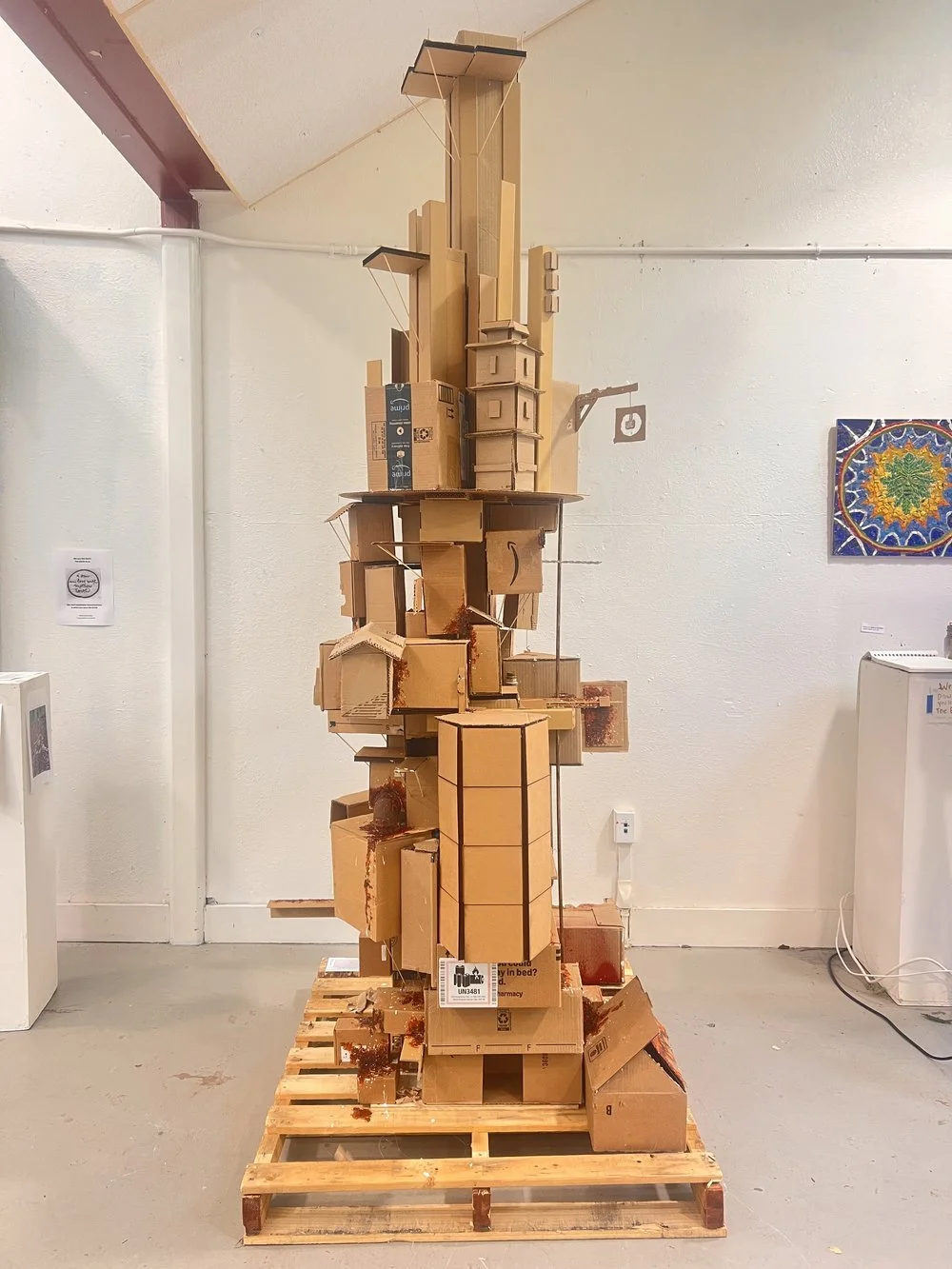 An art sculpture made of stacked cardboard boxes on a wooden pallet in a gallery.