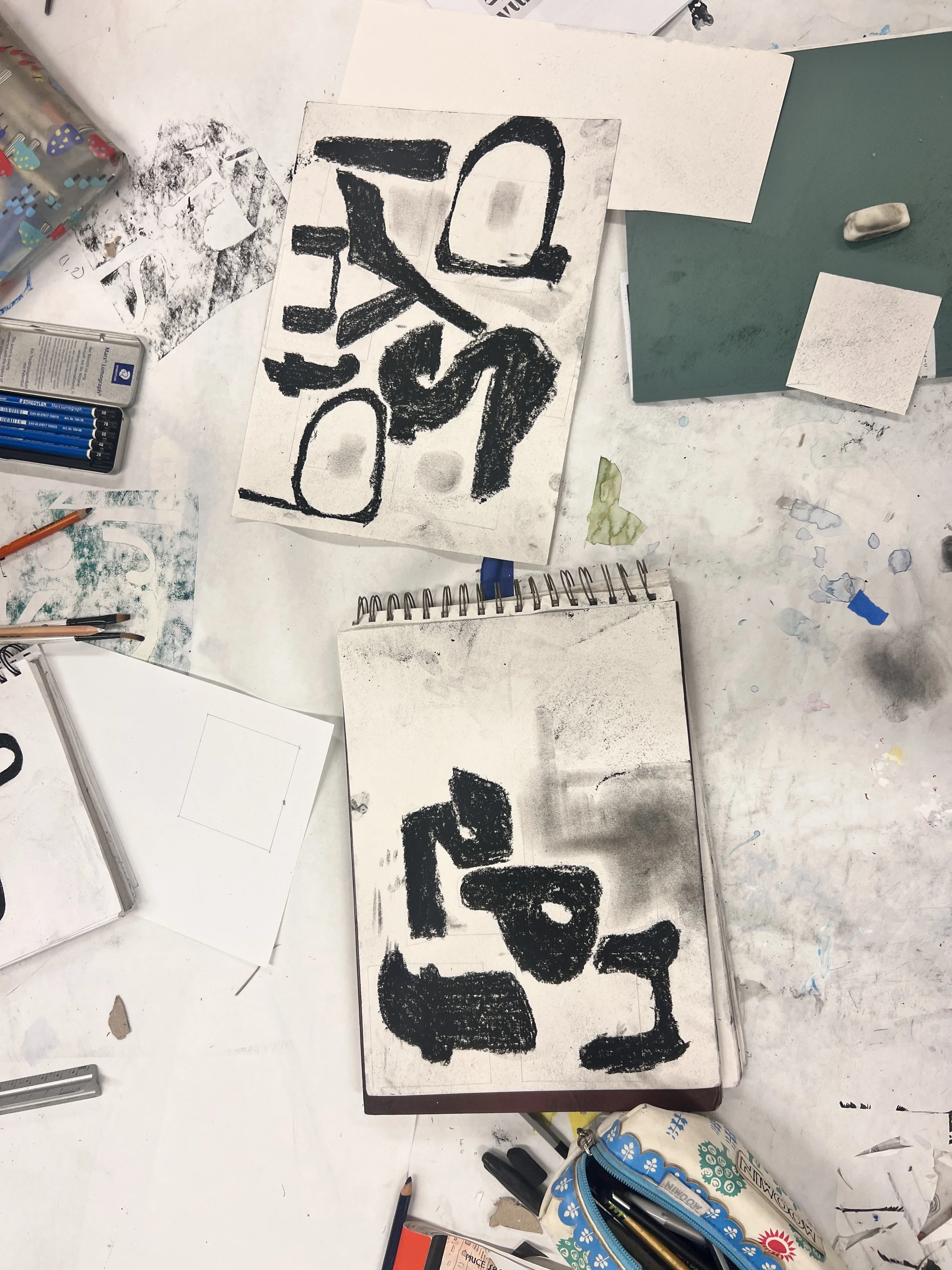 Two sketchbooks featuring abstract, black textured drawings on white pages, surrounded by art supplies including pens, pencils, erasers, paints, and paper on a cluttered table.