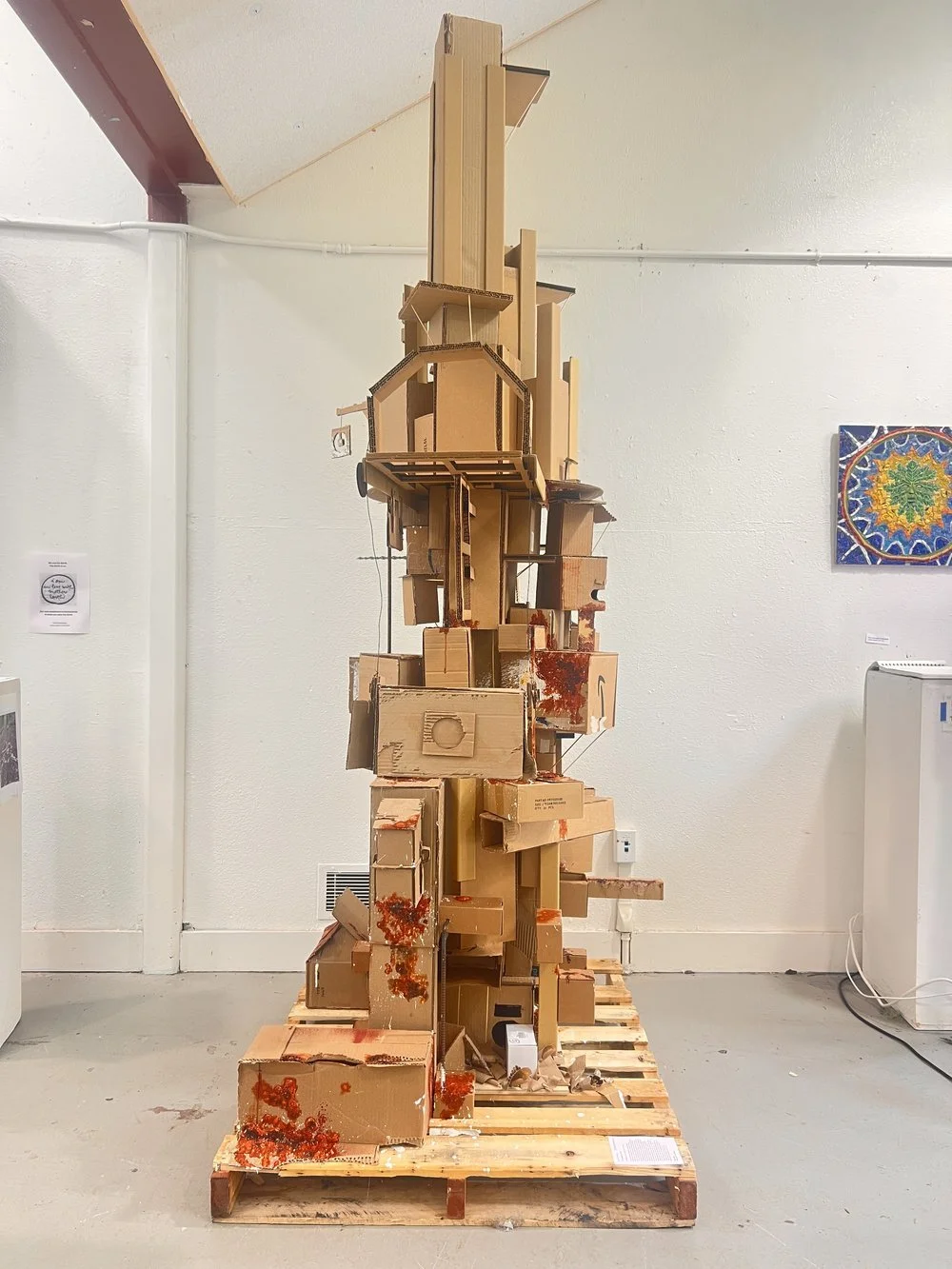 Sculpture made of cardboard boxes and scrap materials, resembling an abstract tower, displayed on a wooden pallet in an art gallery.