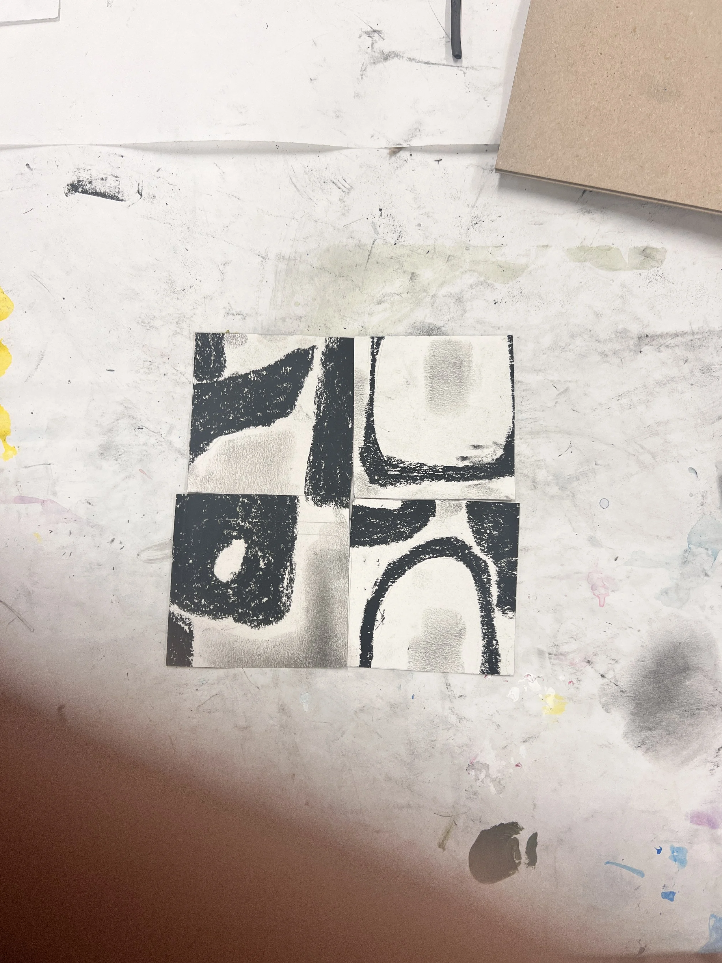 Four black and white artistic prints or stencils on small square tiles, arranged in a 2x2 grid, on a work table with paint and art supplies.