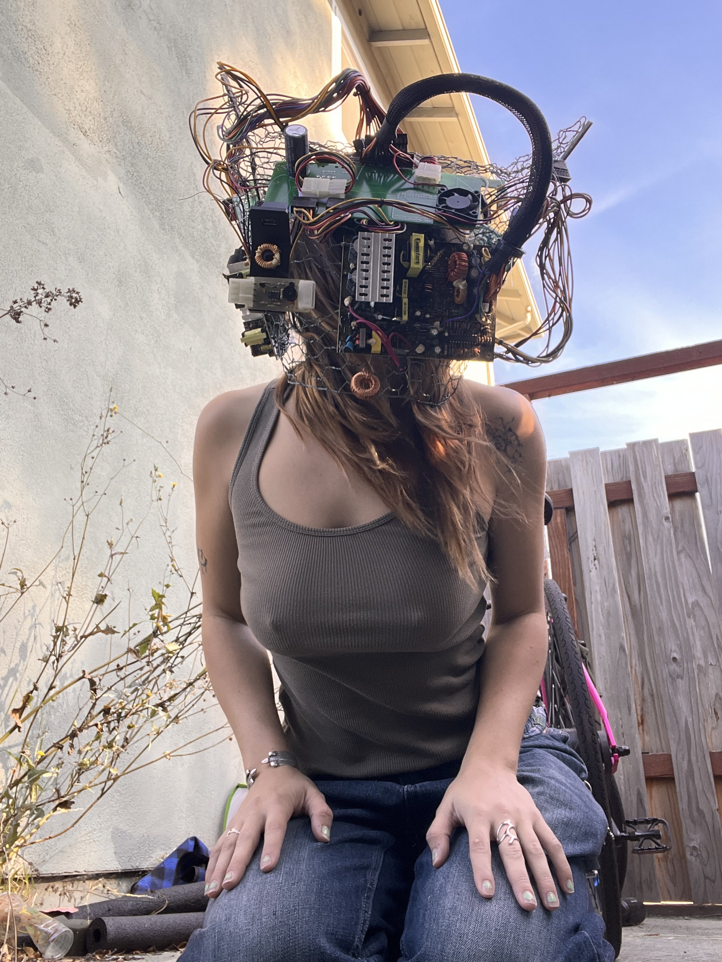 Person kneeling outdoors with a robotic or electronic device on their head, resembling a complex circuit board and wiring. The person has long hair, is wearing a sleeveless top, and is surrounded by a wooden fence and a building with a roof.