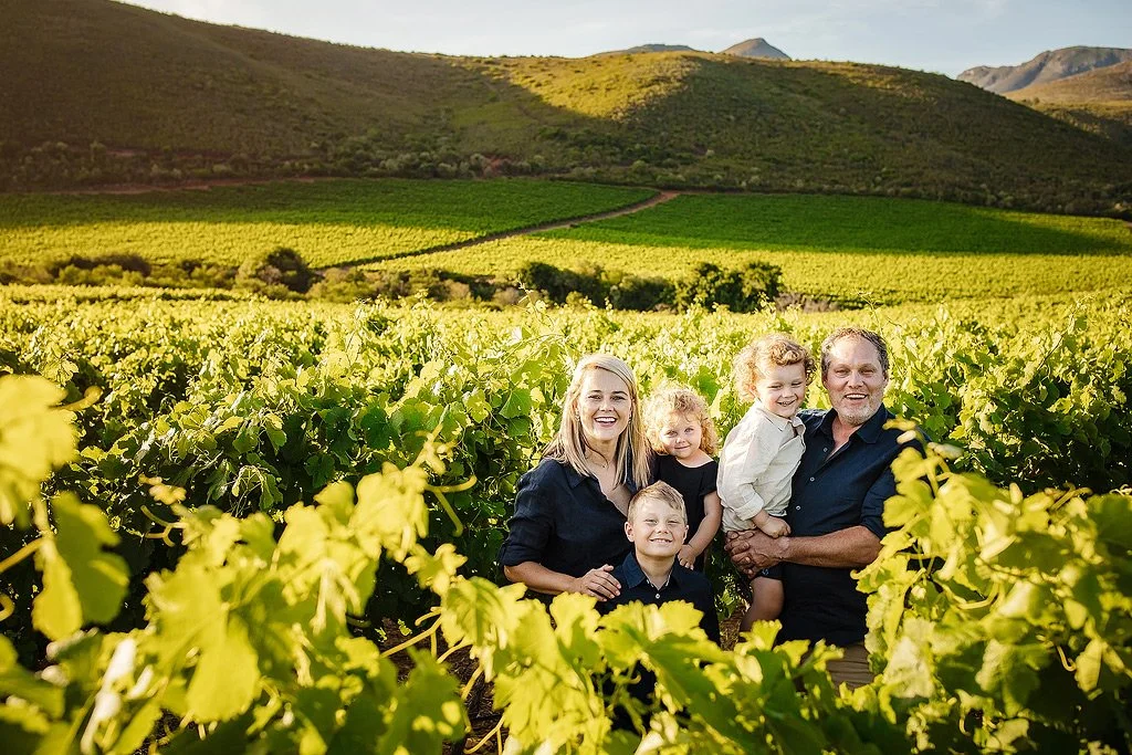 Family of five smiling together in a vineyard with rolling hills in the background during daylight.