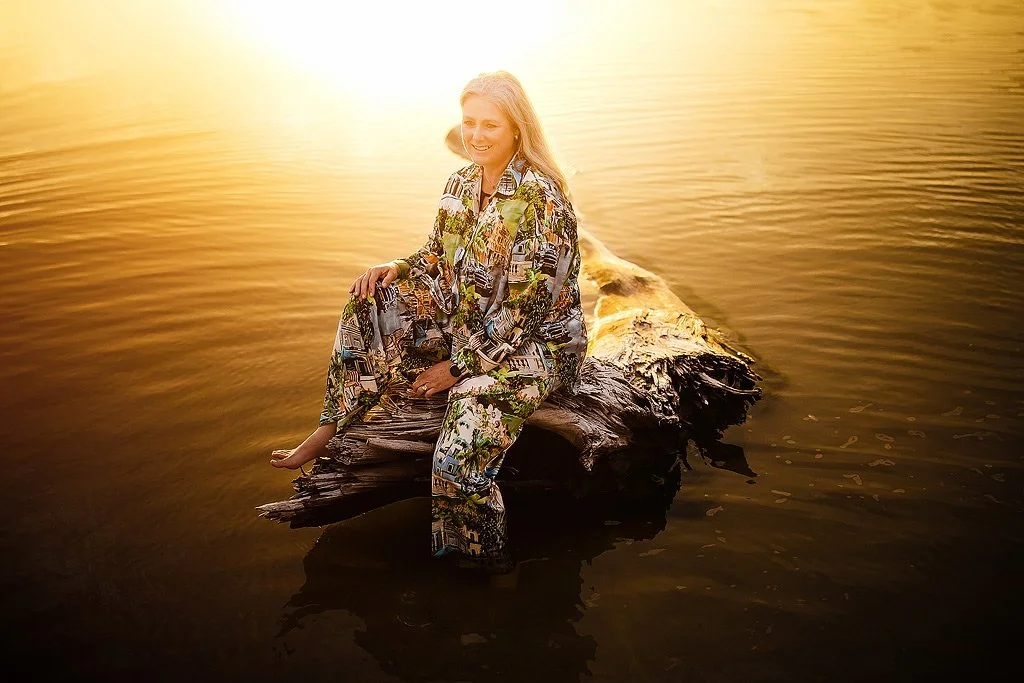 A woman in patterned clothing sitting on a log in the water during sunset.