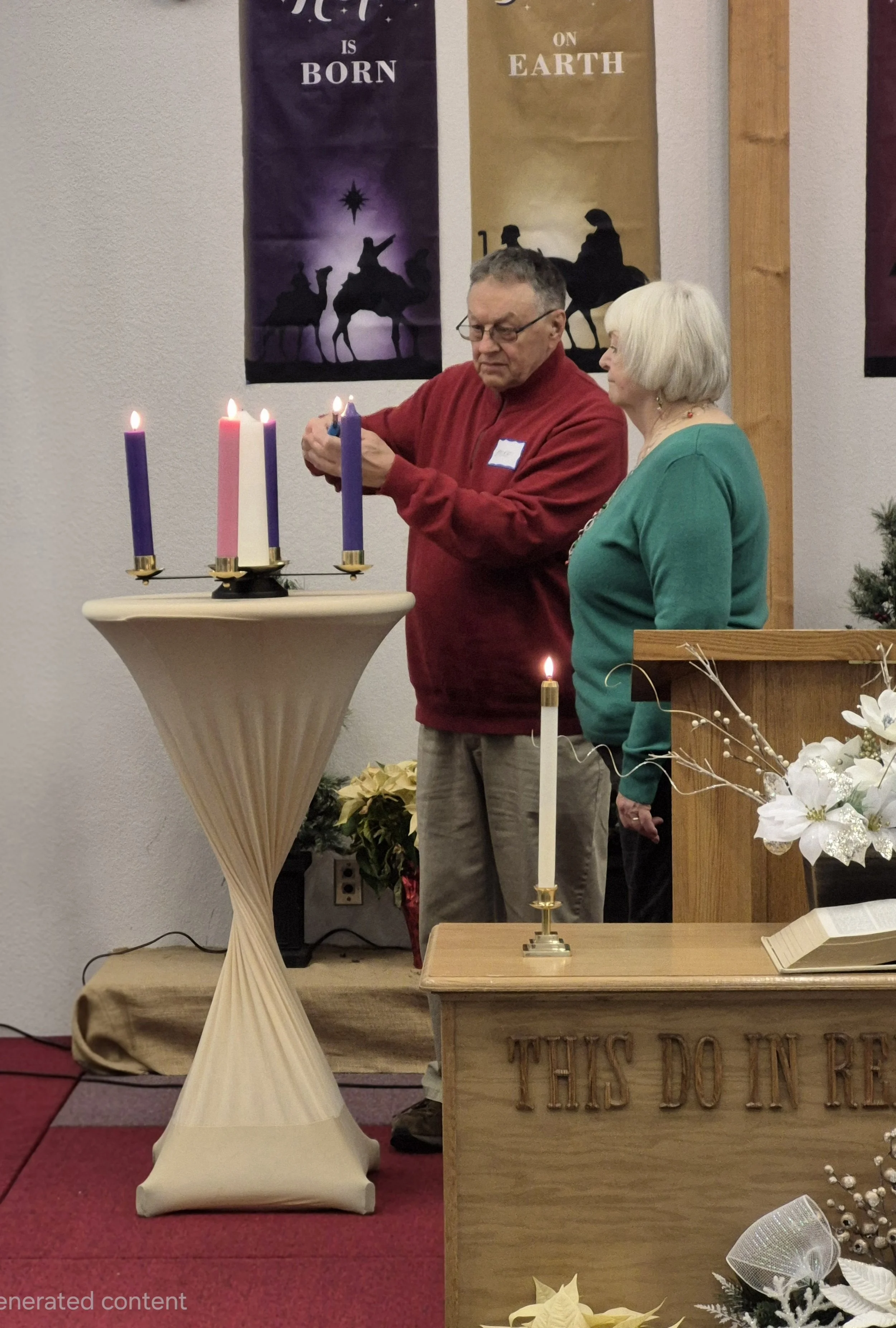 A man and woman standing beside a tall table with four lit candles, a Bible, and Christmas decorations, in a church setting. The man is lighting or adjusting the candles while the woman observes. Banners with biblical themes are hanging on the wall behind them.