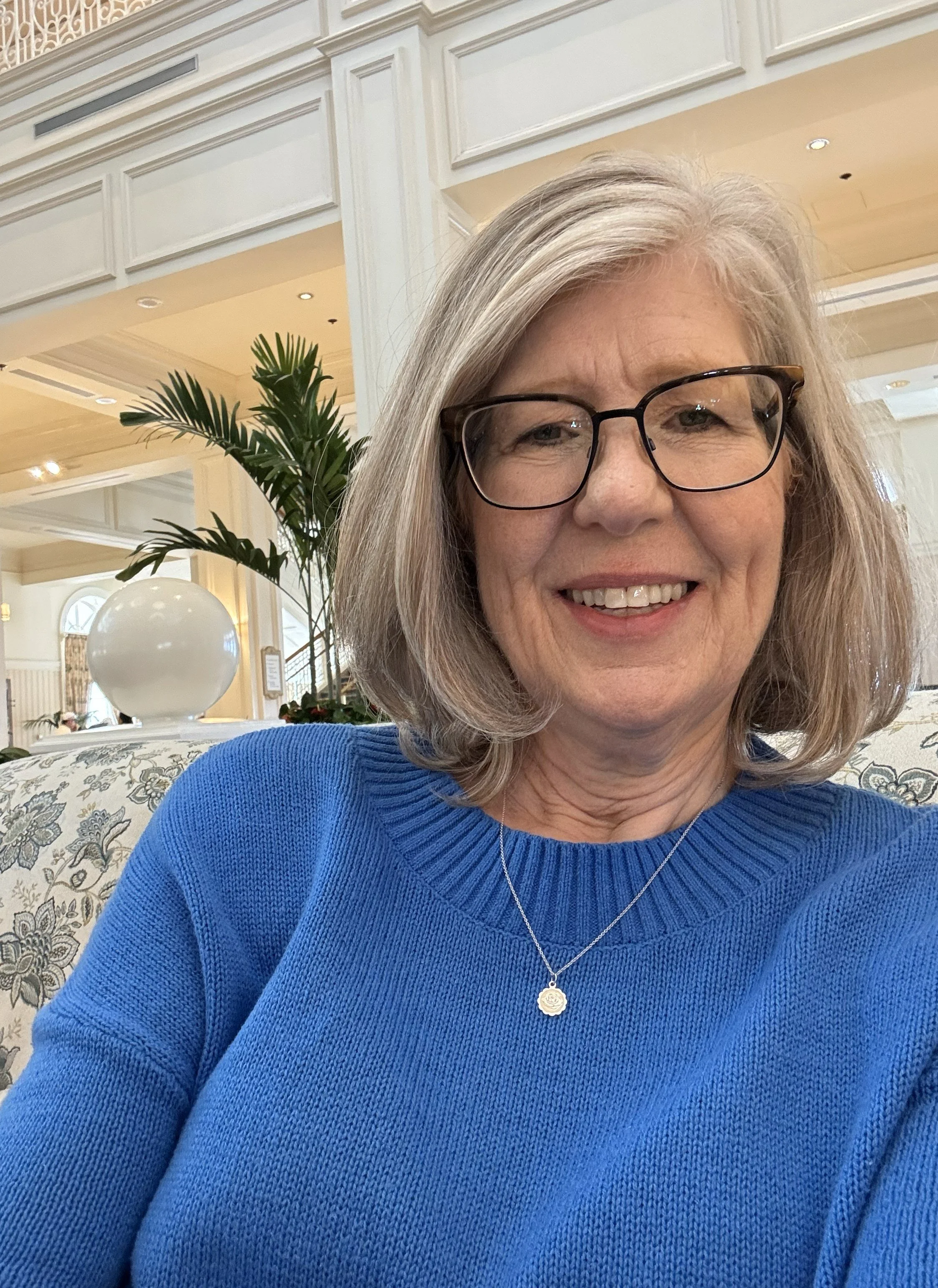 Smiling woman with glasses and gray hair in a hotel lobby, wearing a blue sweater