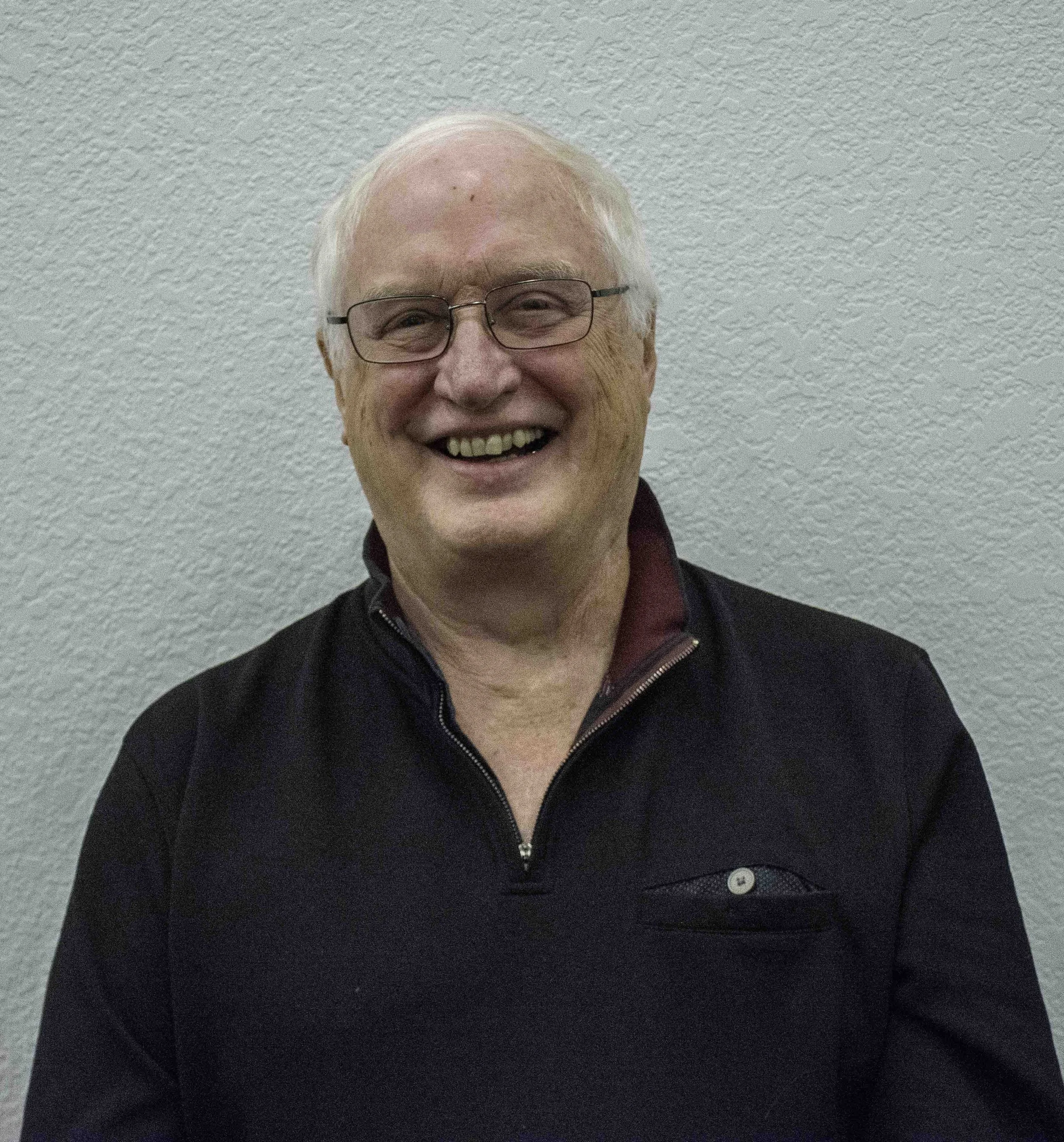 Elderly man wearing a black shirt, smiling against a textured wall.