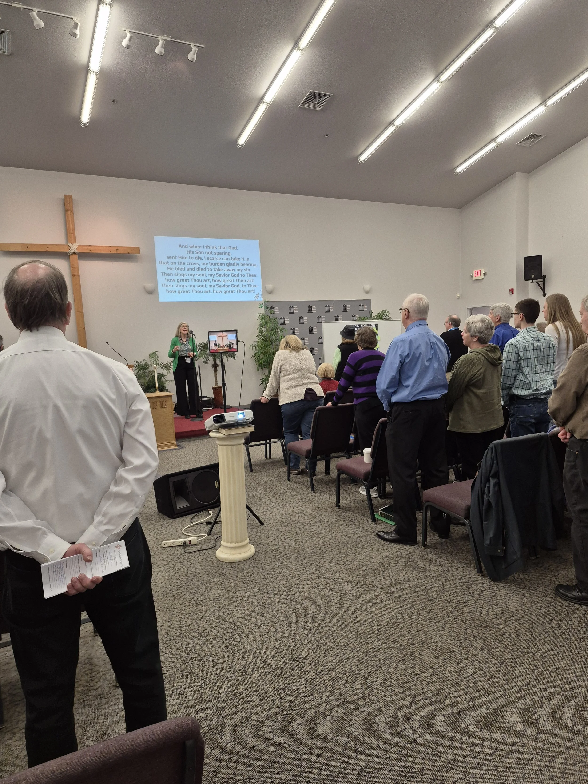 People standing and singing during a church service or gathering in a room with a cross on the wall, a screen displaying lyrics, and a woman leading worship at the front.