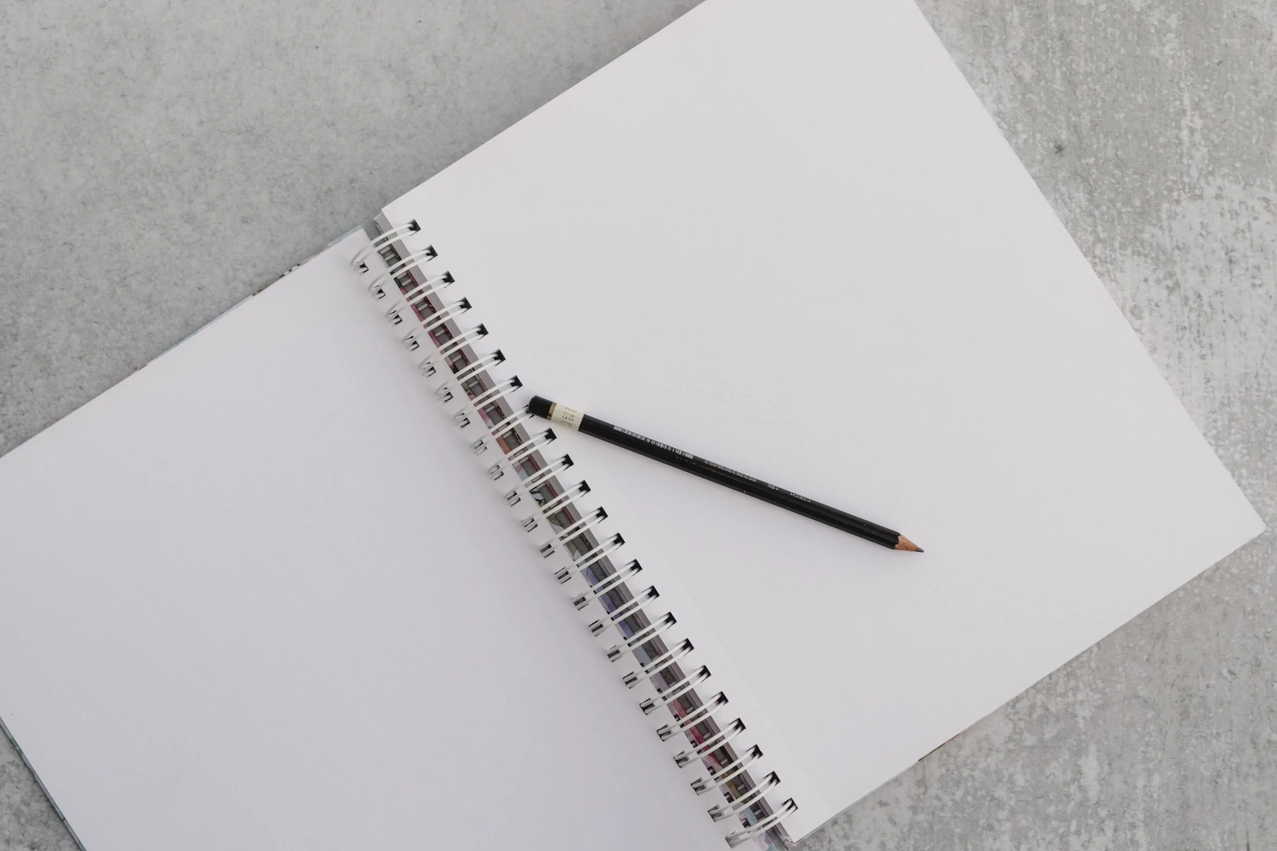 Open spiral notebook with blank white pages and a black pencil lying across the pages on a gray textured surface.