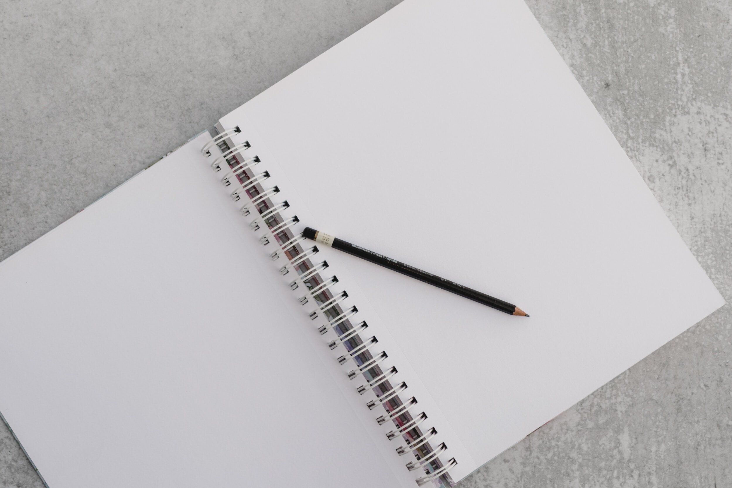 An open spiral-bound notebook with blank white pages on a gray surface, with a black pencil resting diagonally across the center.