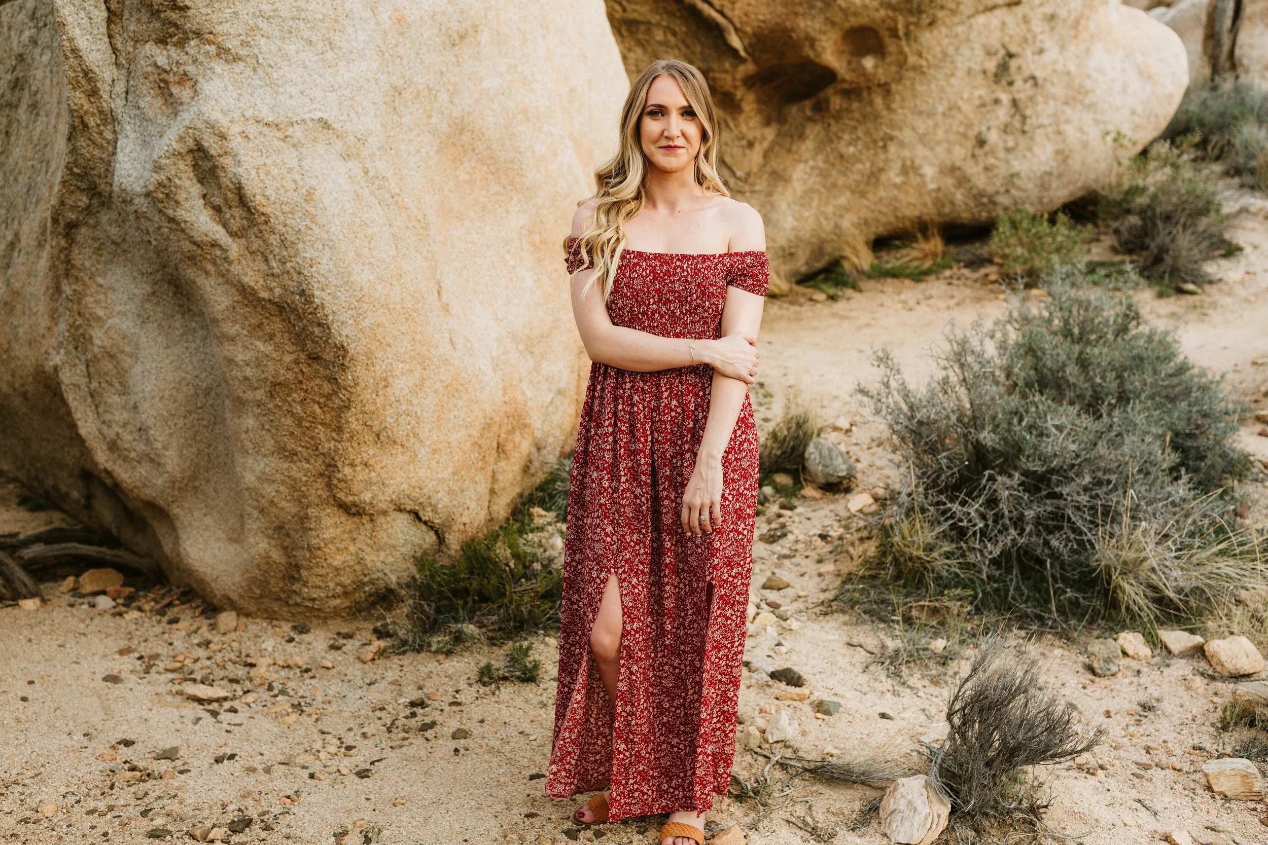 A woman in a red floral off-shoulder dress with a slit standing in a desert-like environment with large rocks and sparse desert plants.