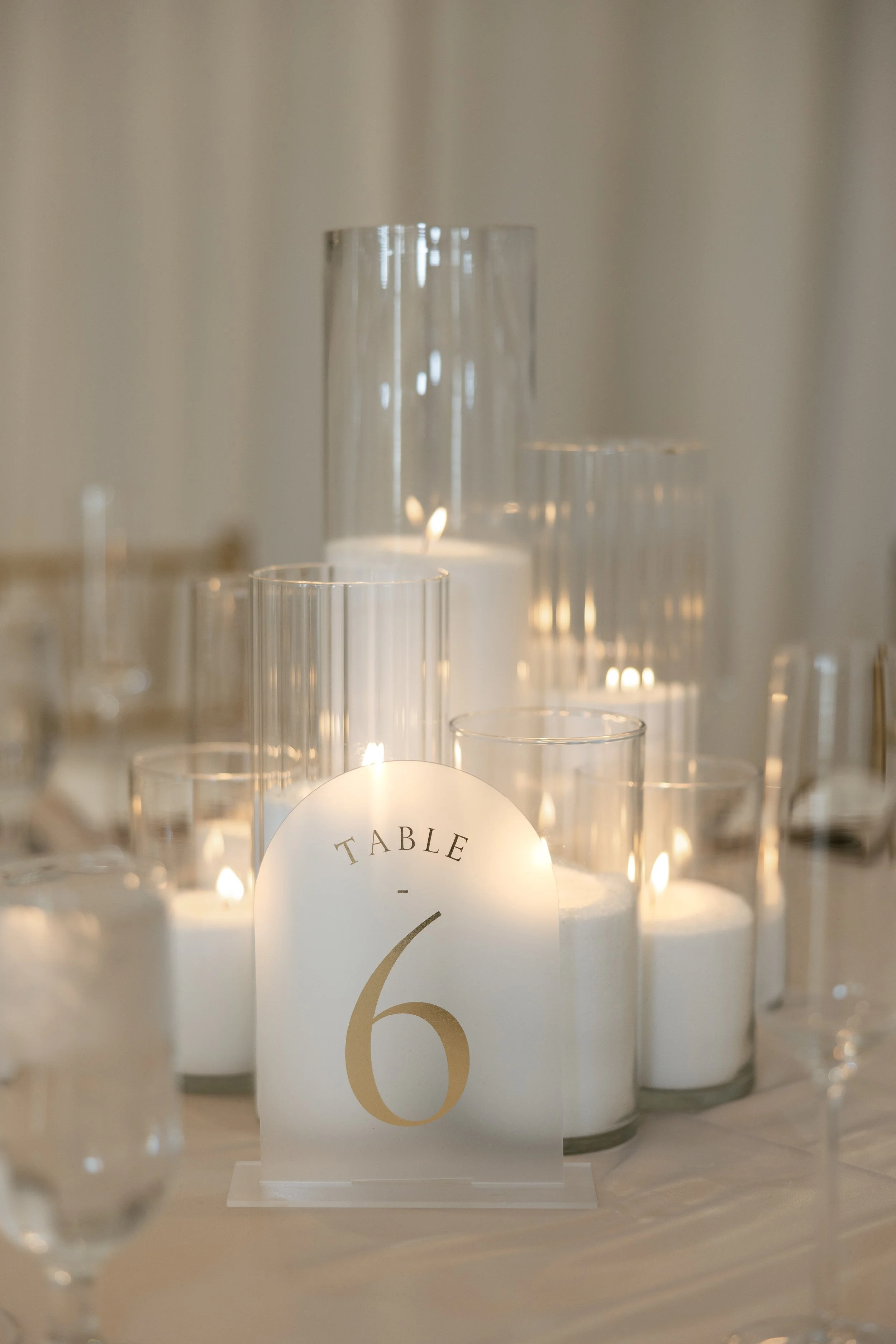 Close-up of a decorative table number 6 sign with glass candle holders and lit candles in the background, on a table set for an event or wedding.