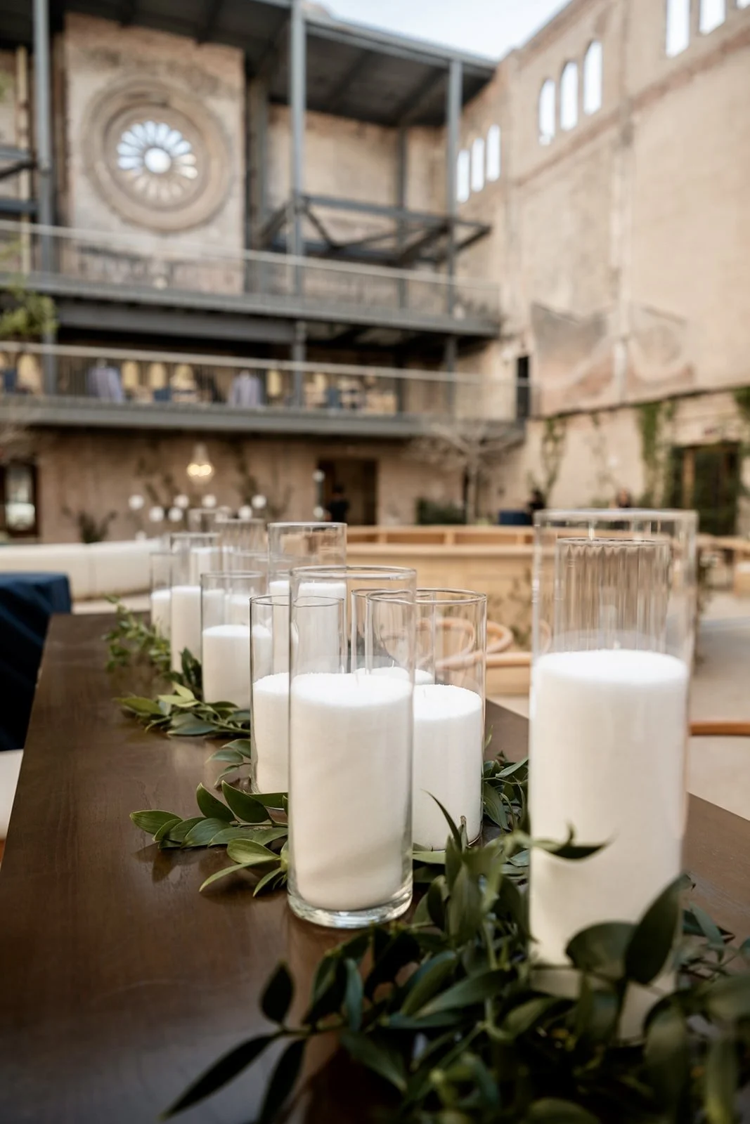 Tall glass candles arranged on a table with leafy greenery in a rustic indoor/outdoor setting, with an exposed brick wall and industrial-style balcony in the background.