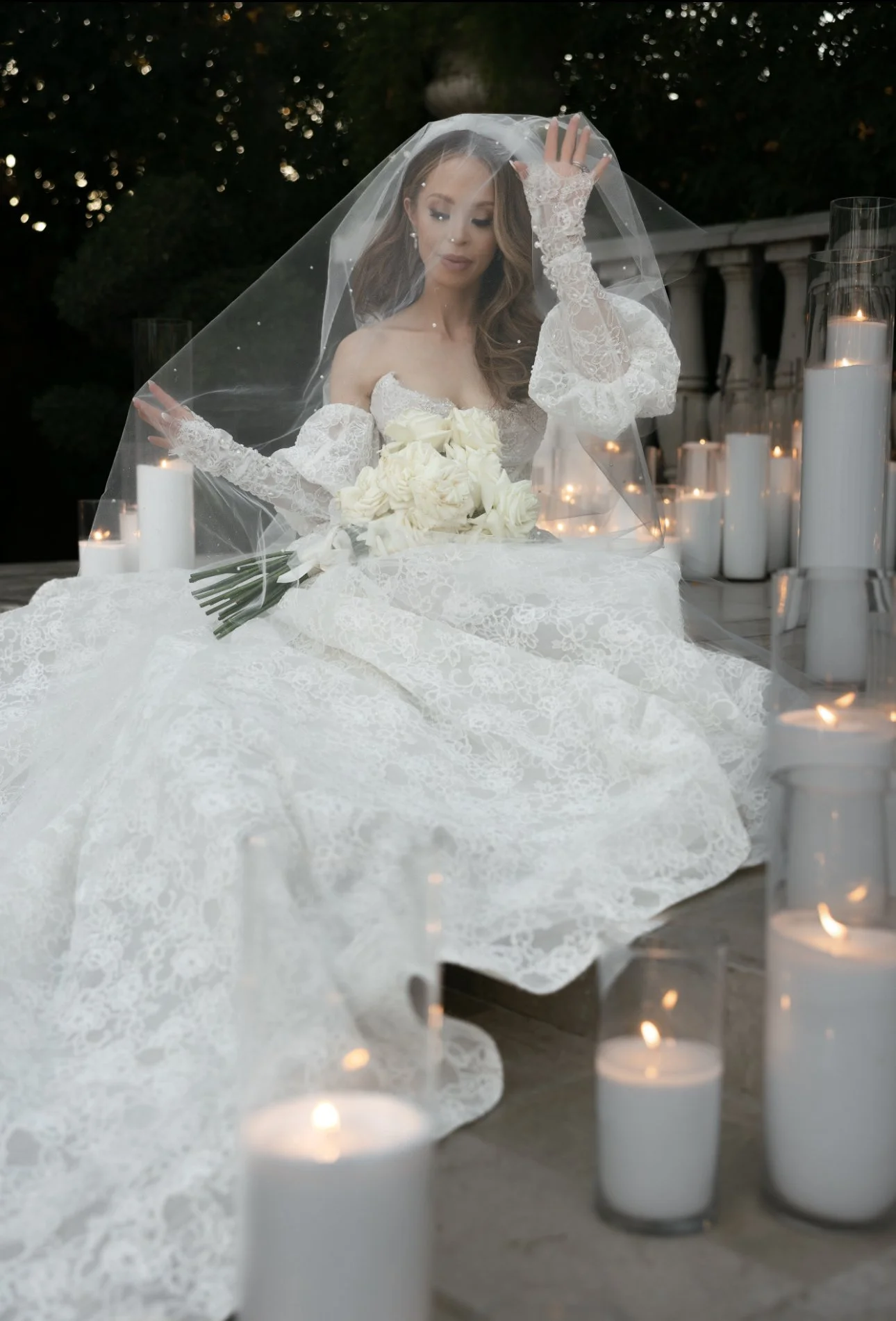 A bride in a lace wedding dress and veil holding a bouquet of white roses, sitting outdoors surrounded by lit pearl sand candle candles.