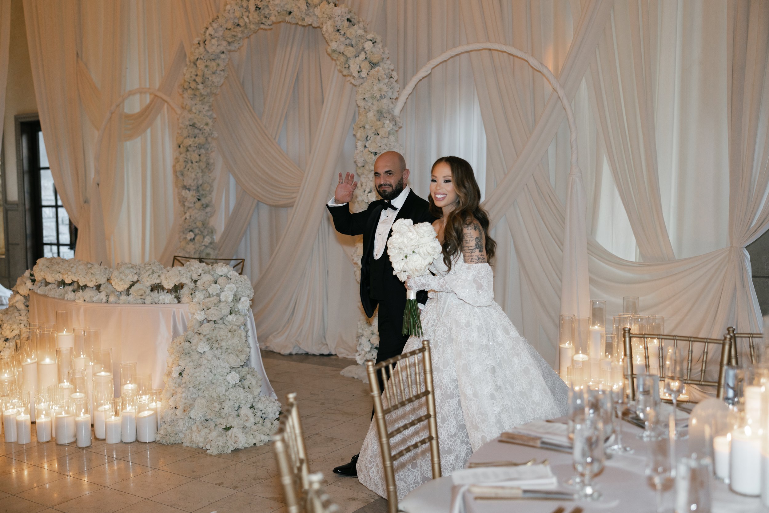 A newly married couple at their wedding reception, standing in front of a decorated backdrop with white drapes and floral arrangements. The groom is dressed in a black tuxedo with a black bow tie, and the bride is in a white lace wedding gown holding