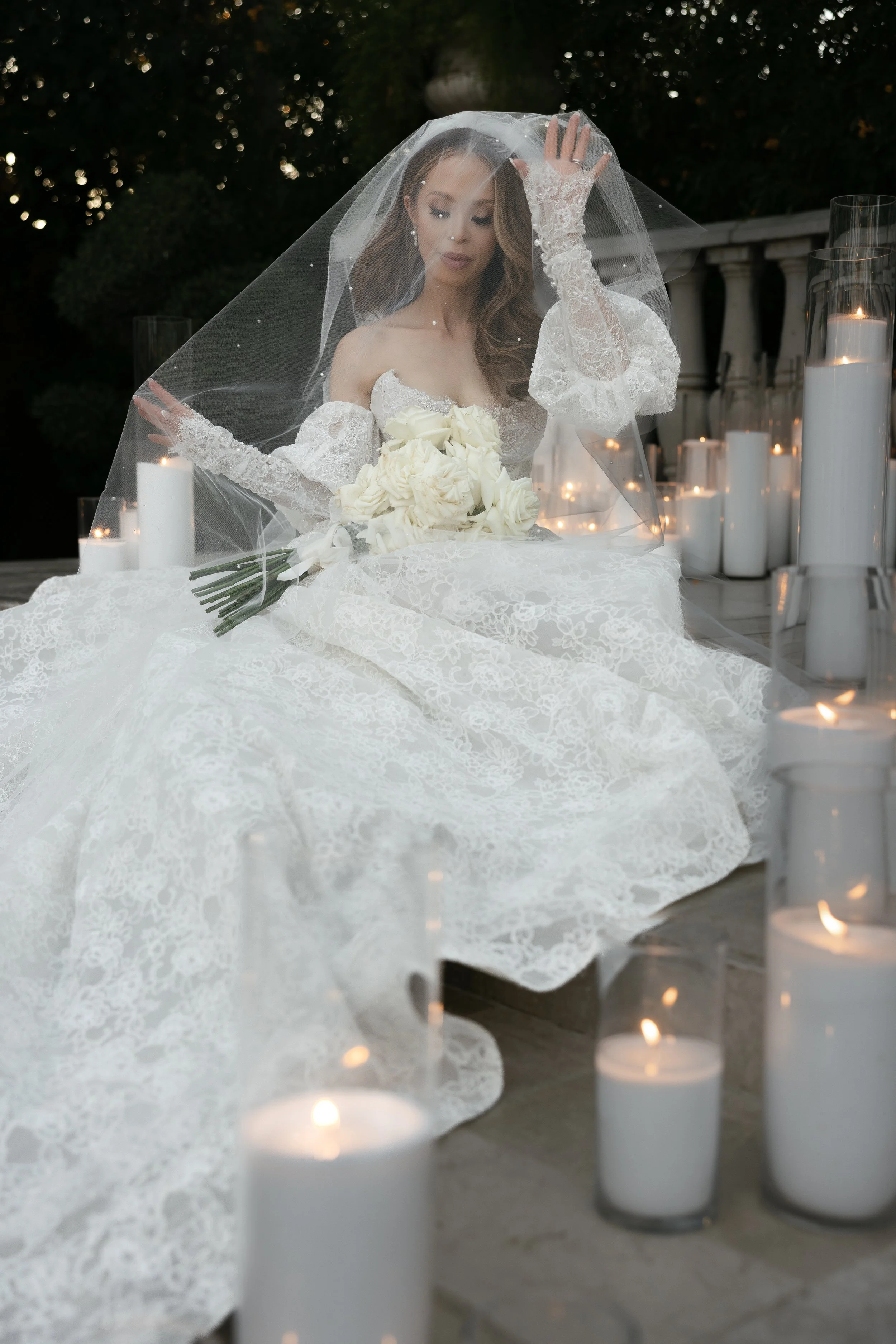 A bride in a white lace wedding gown and veil sitting outdoors at night, holding a bouquet of white roses, surrounded by lit white pearl sand candles.