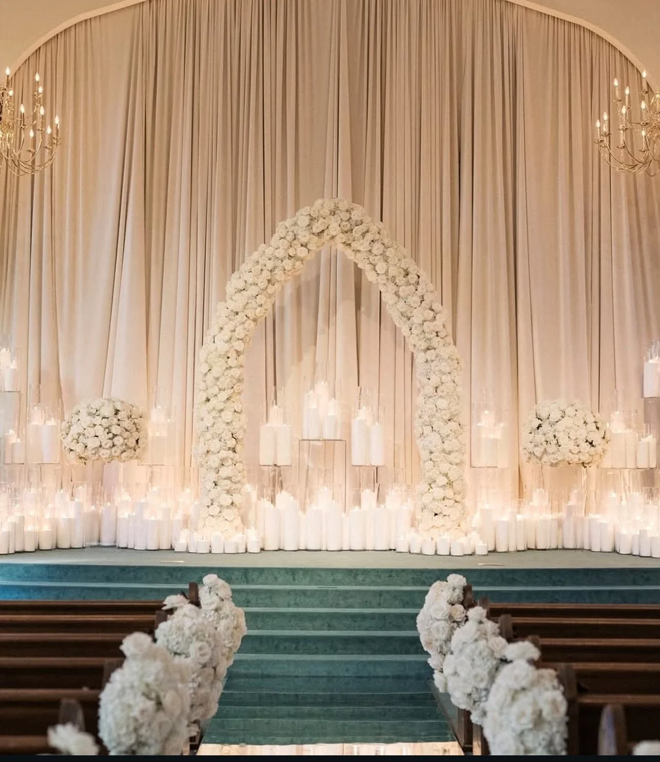 Elegant wedding arch decorated with white flowers, with draped beige curtains and soft lighting, leading to an aisle lined with white floral arrangements.