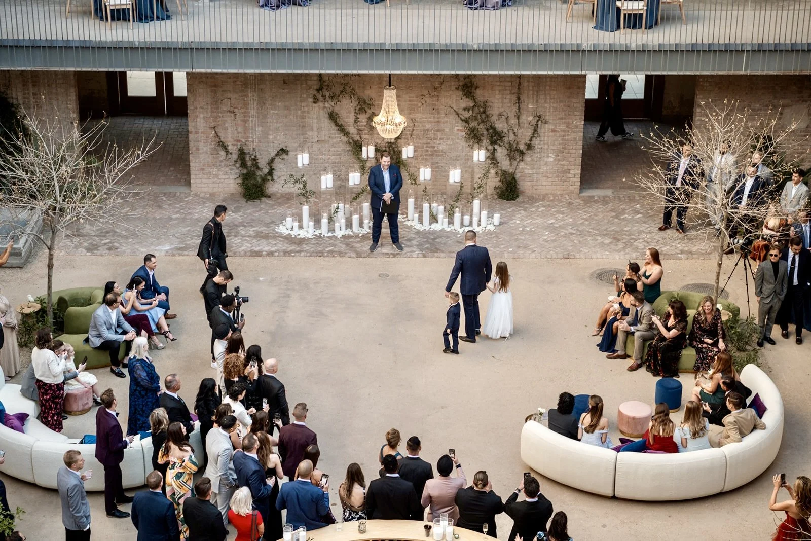 A wedding ceremony is taking place in an indoor venue with a large crowd of guests. The bride, wearing a white dress, and the groom, in a dark suit, are standing in front of a person officiating. There are candles and greenery decorations behind them