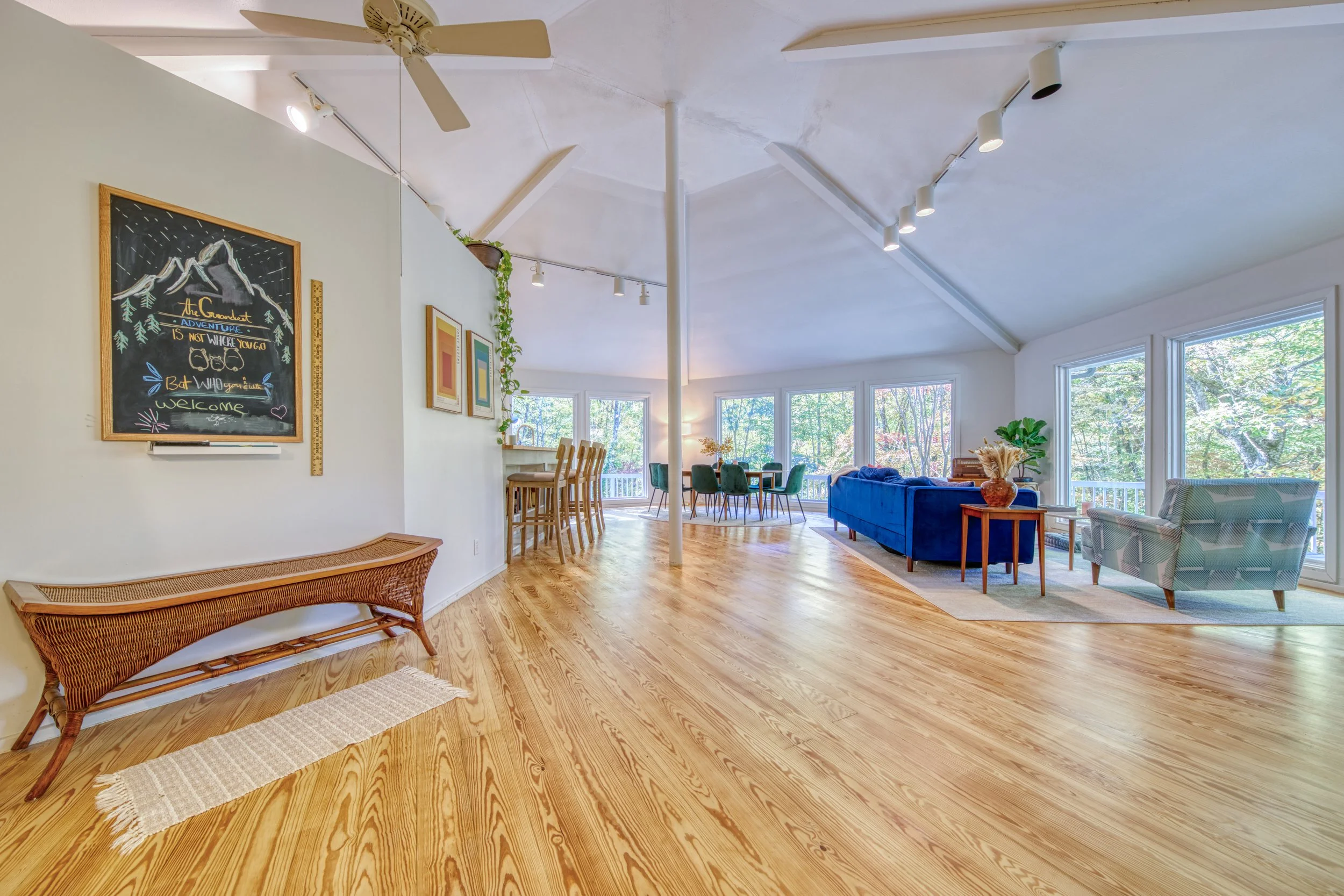 Open-concept living and dining area with large windows, wooden flooring, a blue velvet sofa, green chairs, wall art, and indoor plants.