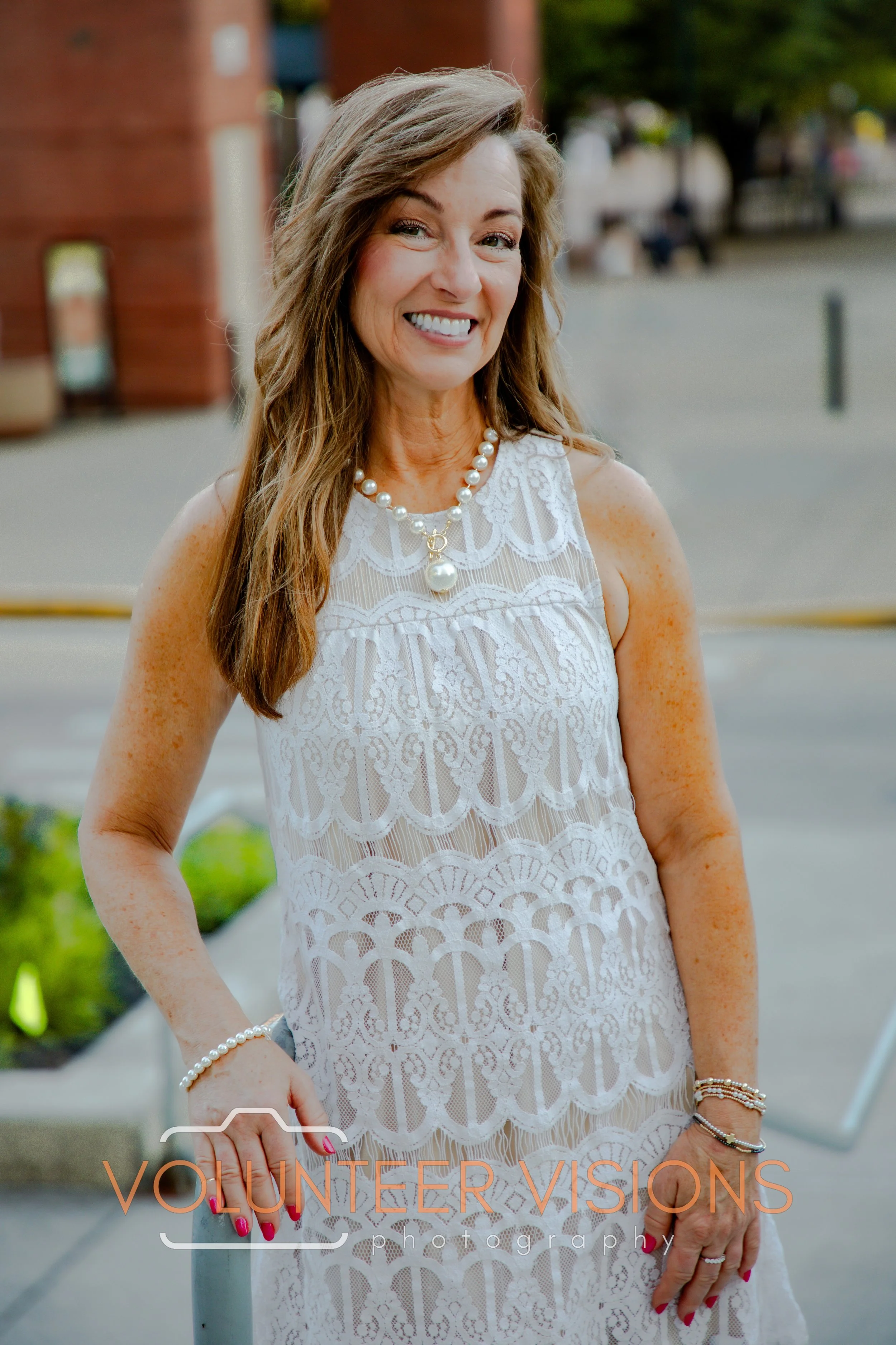 A woman with long hair smiling, wearing a white lace dress and pearl jewelry, standing outdoors with trees and buildings in the background.
