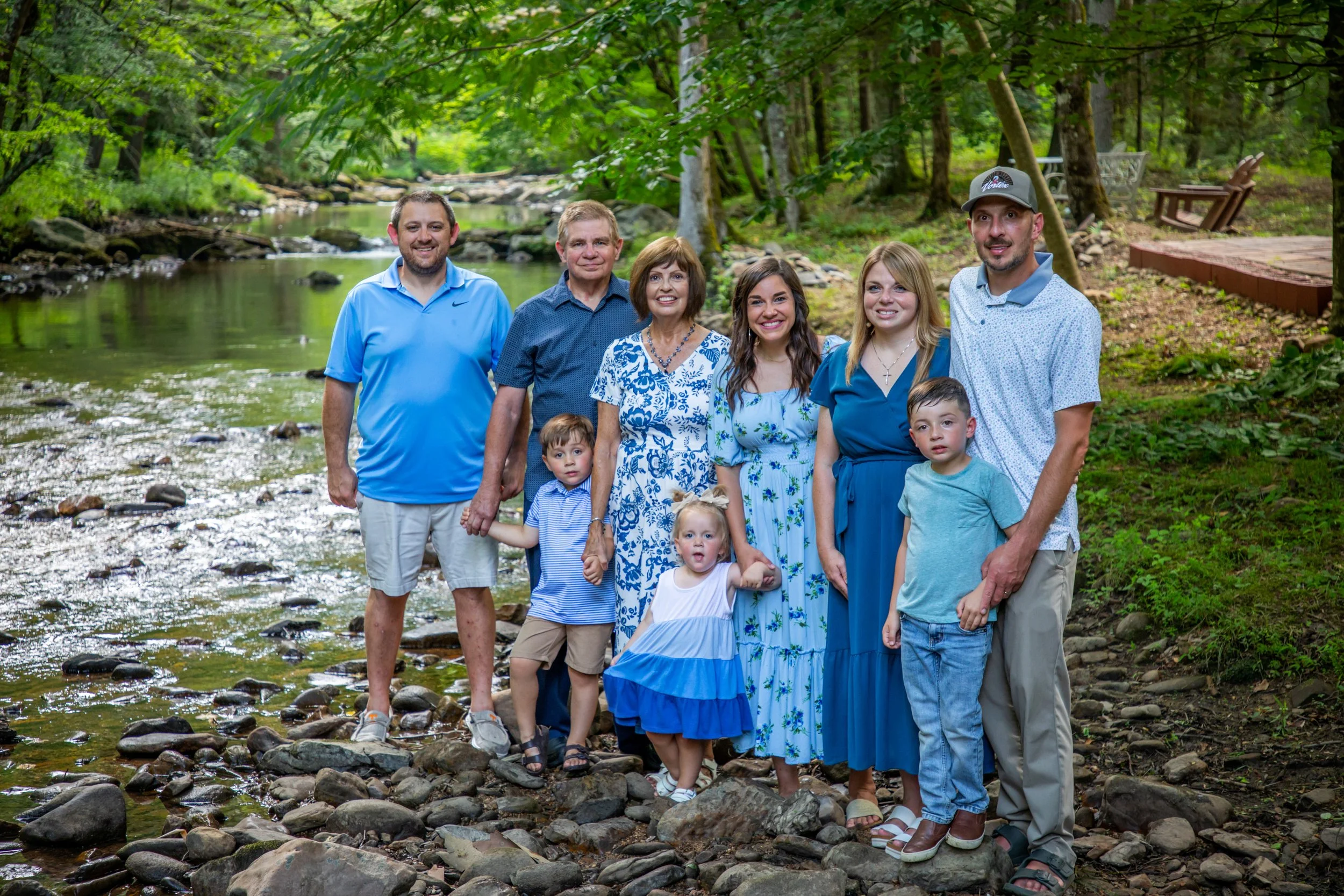 A family of nine people, including adults and children, standing on rocks in a shallow creek in a lush, green forest, smiling at the camera.