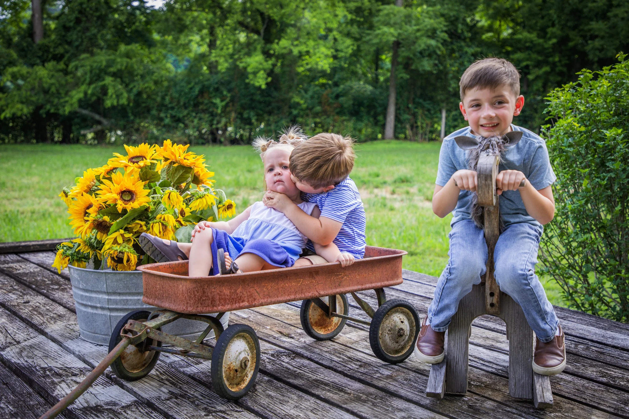 Three young children outdoors with two in a wagon and one sitting on a wooden bench, wearing casual clothes and nature-themed accessories, surrounded by greenery and sunflowers.