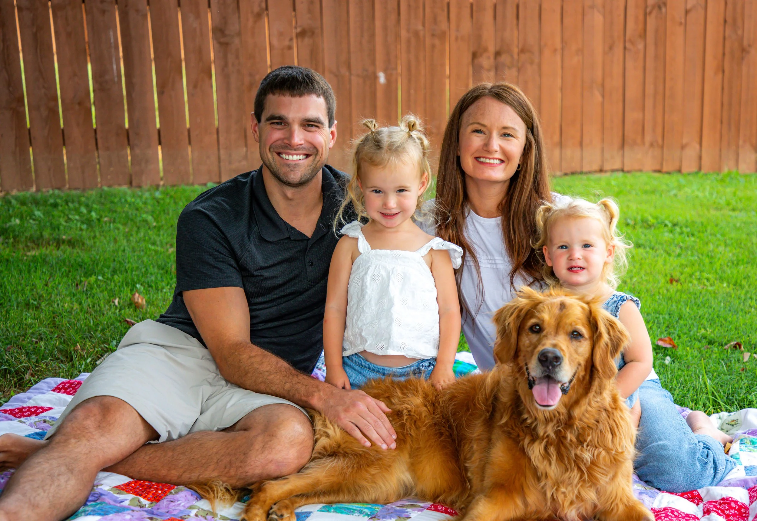 A family of four with two young girls and two adults, sitting outside on a colorful quilt in a backyard, with a large brown dog. The background features a wooden fence and green grass.