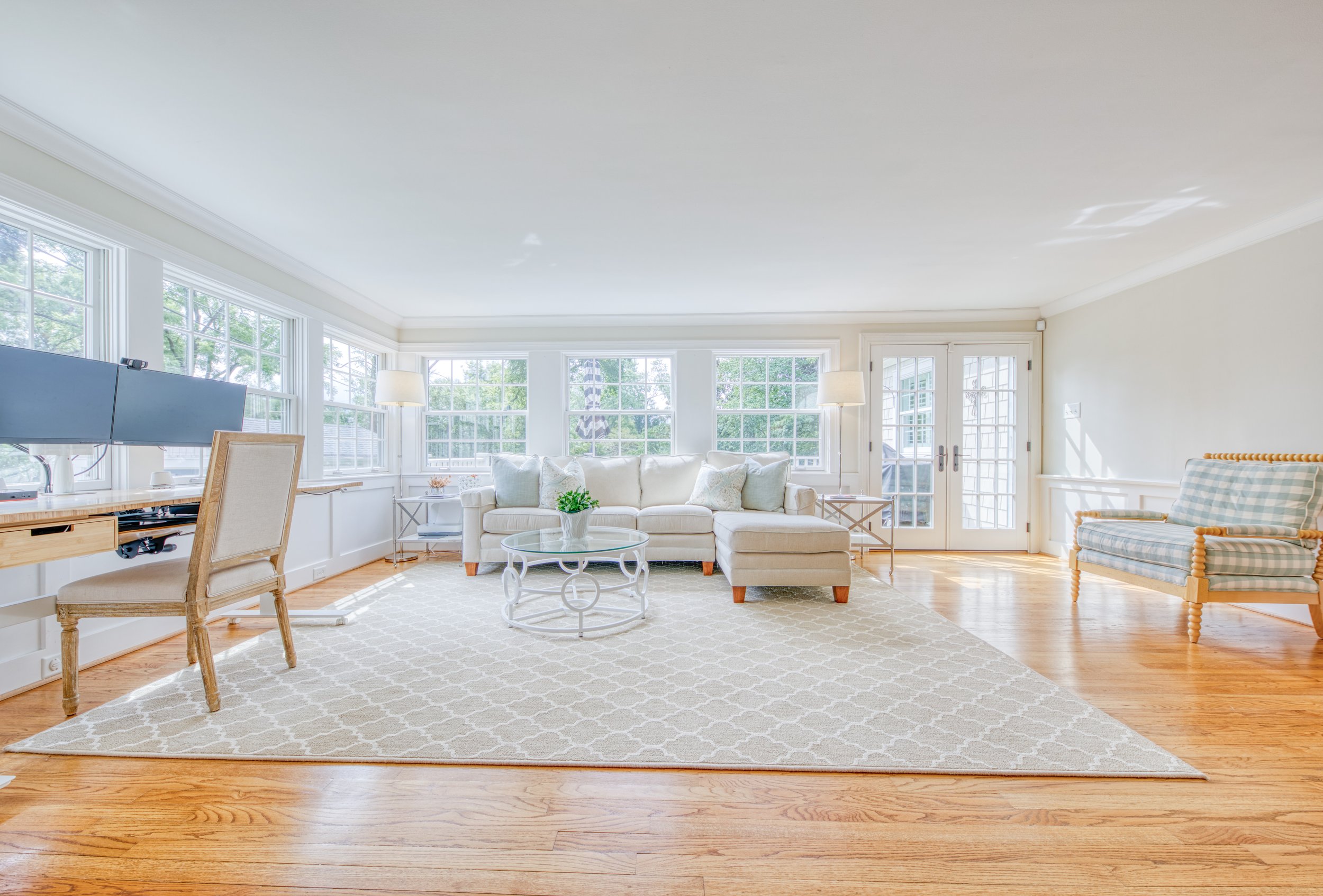 Bright living room with large windows, white sofa, plaid armchair, coffee table, and area rug with natural wood flooring.