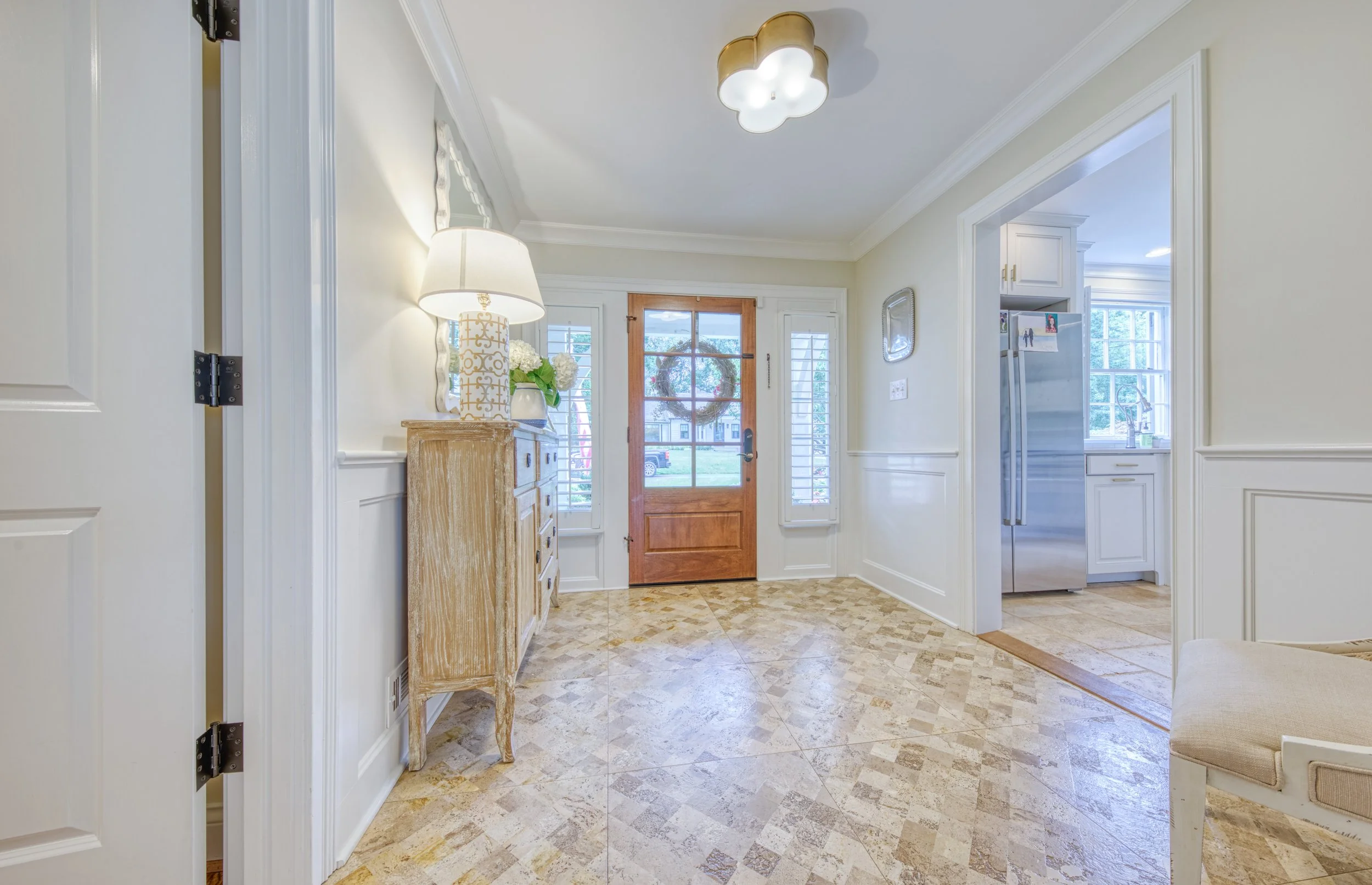 Entryway with front door, a wooden sideboard with a white lamp and flowers, and kitchen visible through an open doorway.