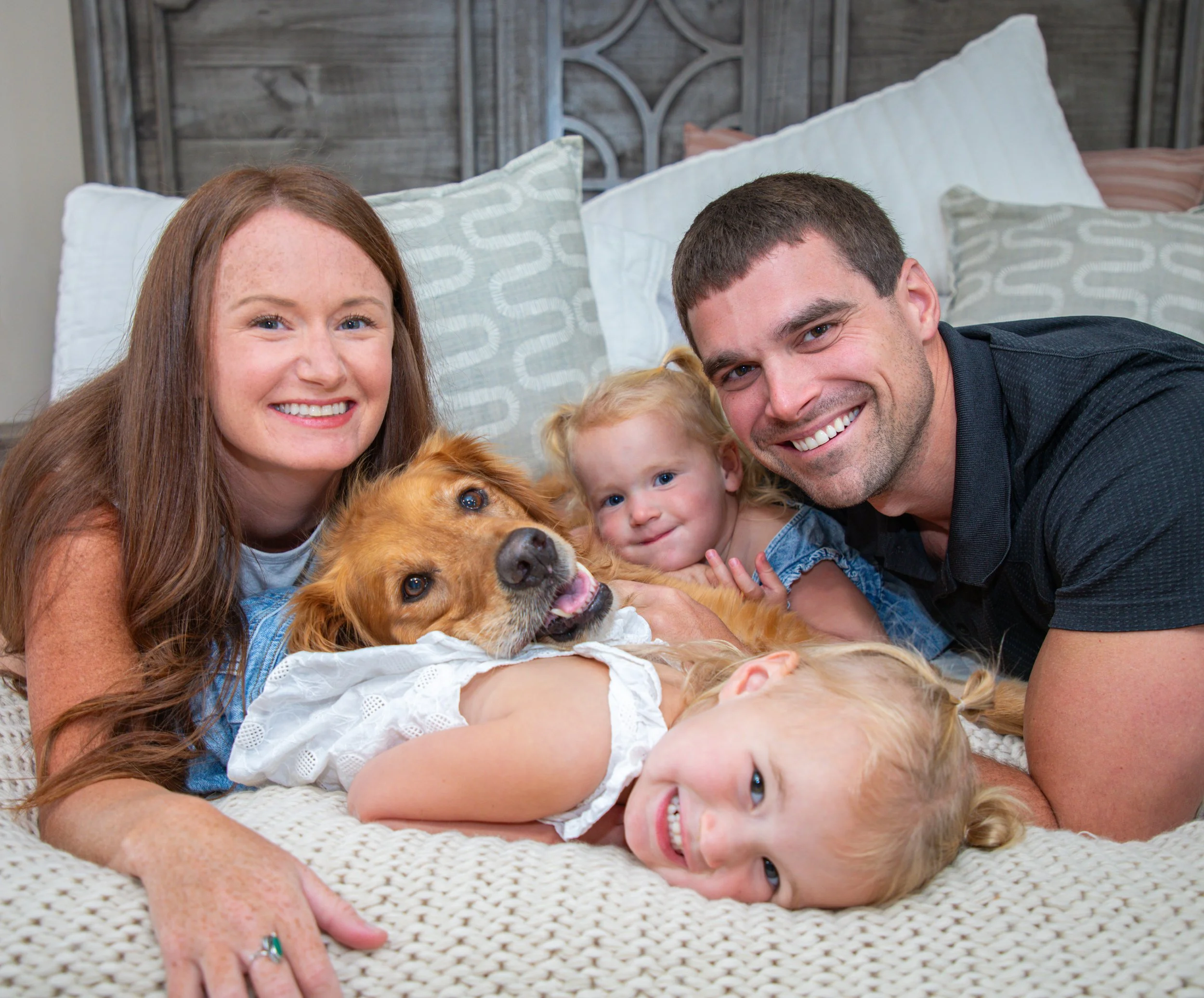 A smiling family lying on a bed with their dog, a young girl, and two boys, all close together with a rustic wooden headboard in the background.