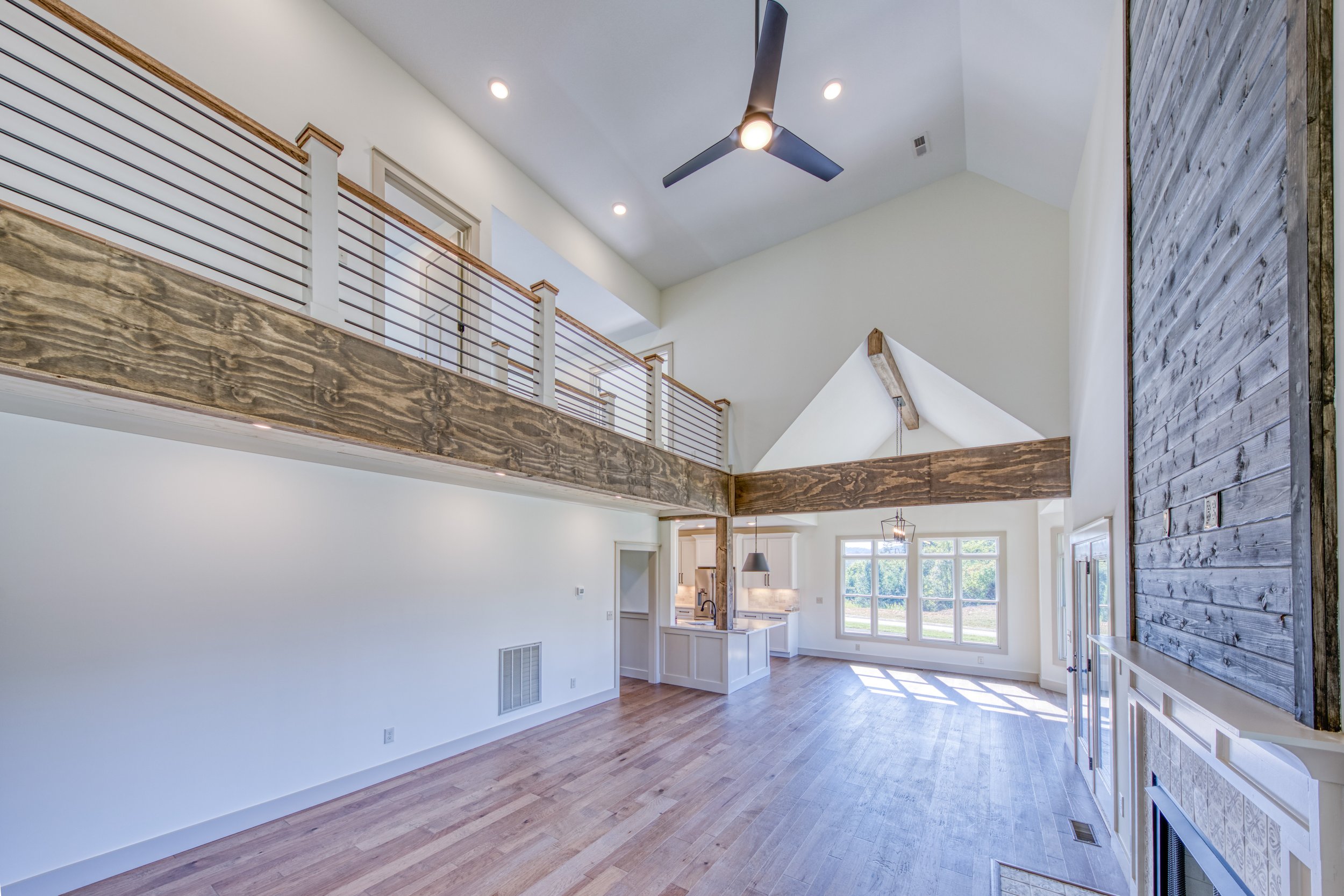 Open-concept living space with vaulted ceiling, large windows, exposed wooden beams, modern ceiling fan, and hardwood floors.