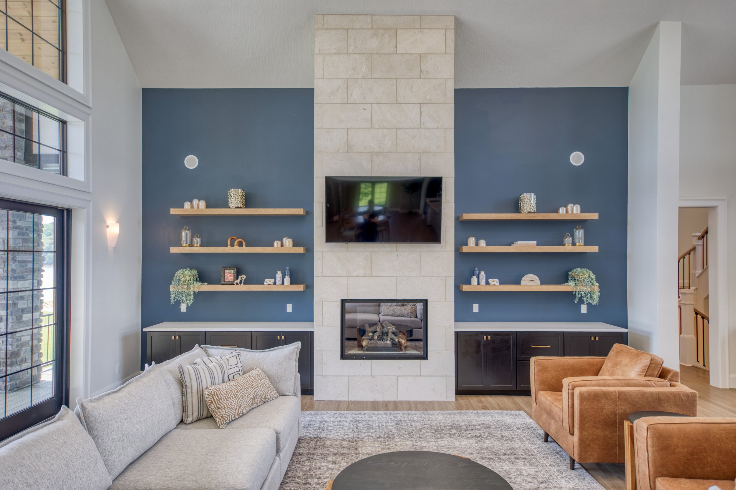 A living room with a beige sofa with patterned pillows, a brown armchair, a rug, and a modern fireplace with a mounted TV above it. There are three floating shelves decorated with candles, vases, and small decor items, against a blue accent wall with