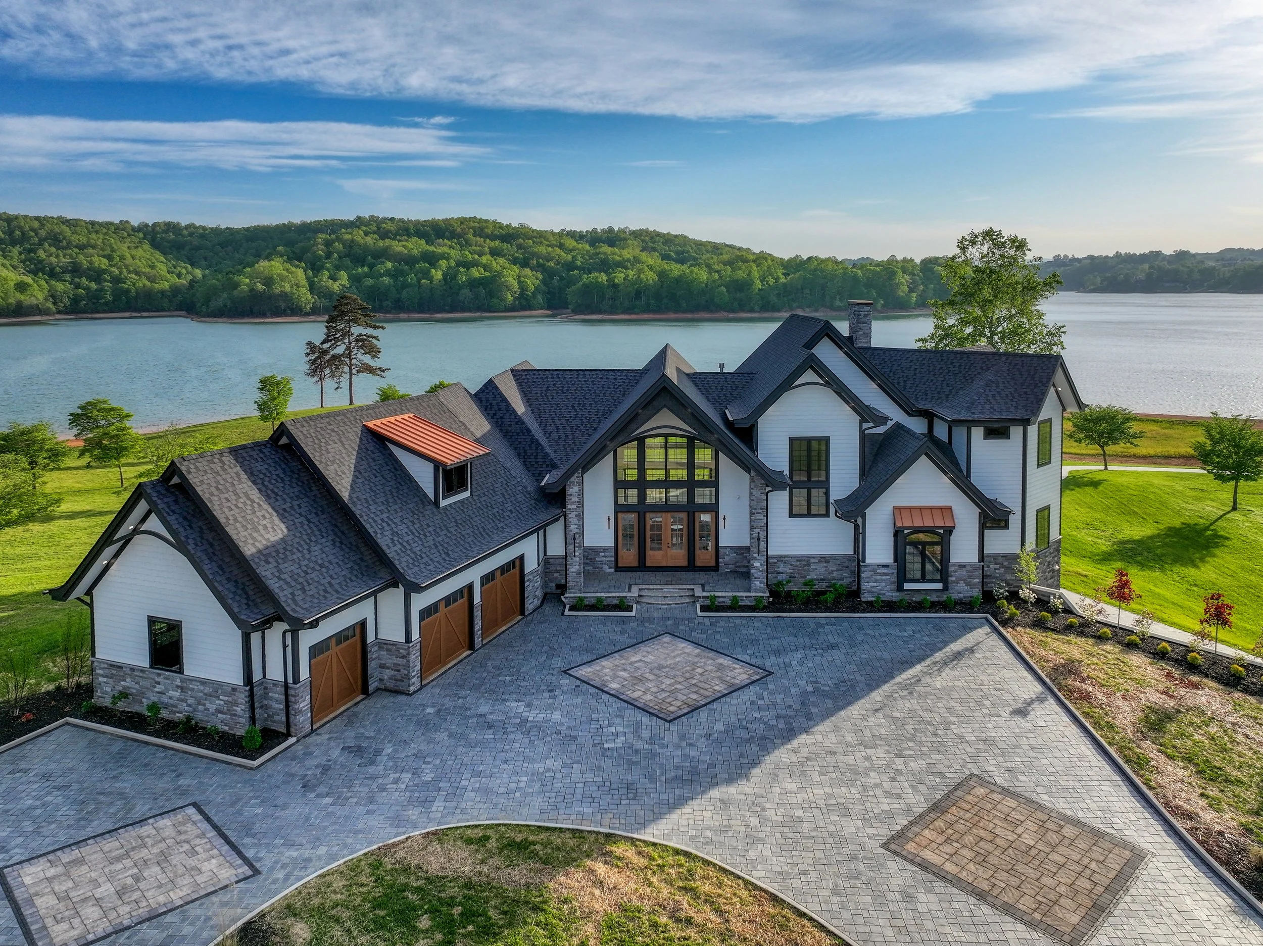 A large modern house with a gray shingled roof, white exterior walls, extensive windows, and a stone chimney, situated by a lake with green hills and a partly cloudy sky in the background.