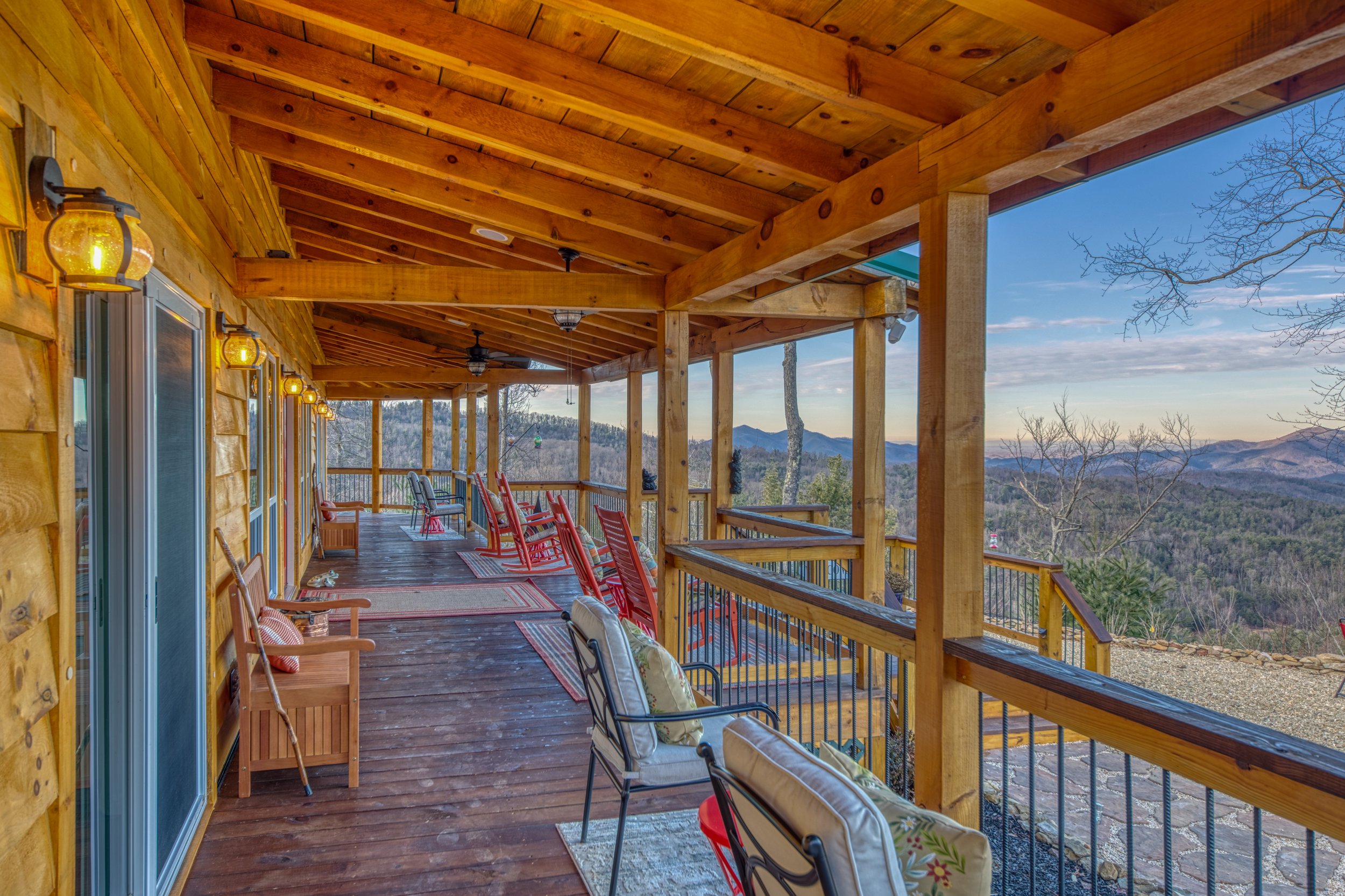 Long porch or patio with wooden flooring and railing, decorated with chairs and rugs, overlooking a scenic mountain view with trees and a blue sky.