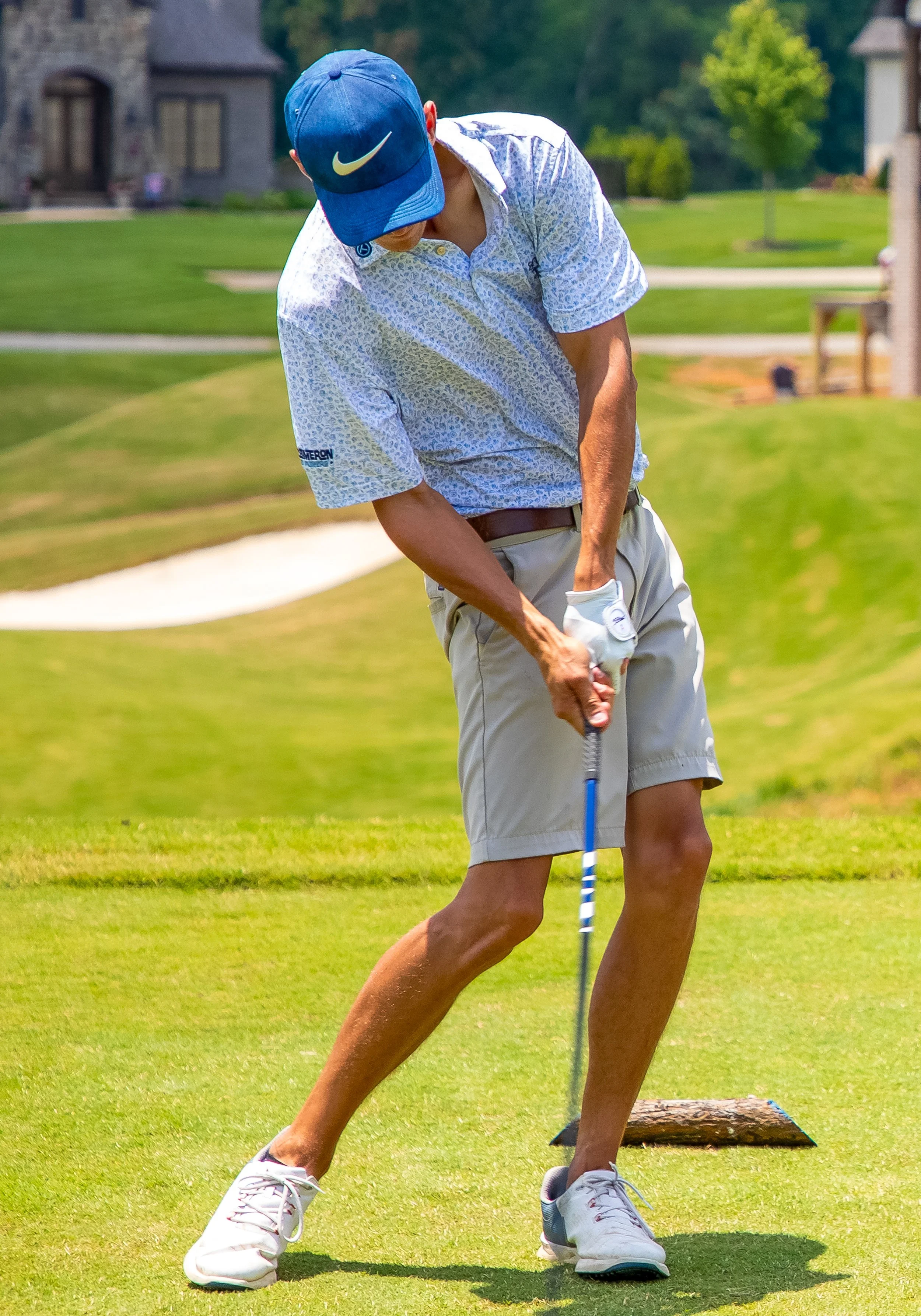 A man playing golf on a lush green course, wearing a blue Nike cap, a patterned short-sleeve shirt, beige shorts, and white golf shoes, preparing to hit the golf ball.