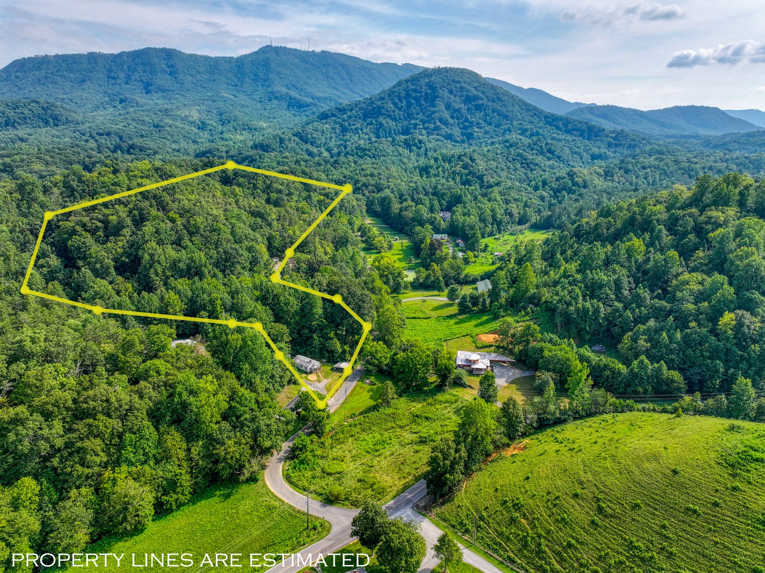 Aerial view of a lush, green mountainous landscape with a yellow property boundary outline surrounding a wooded area and some structures, with distant hills and a partly cloudy sky in the background.