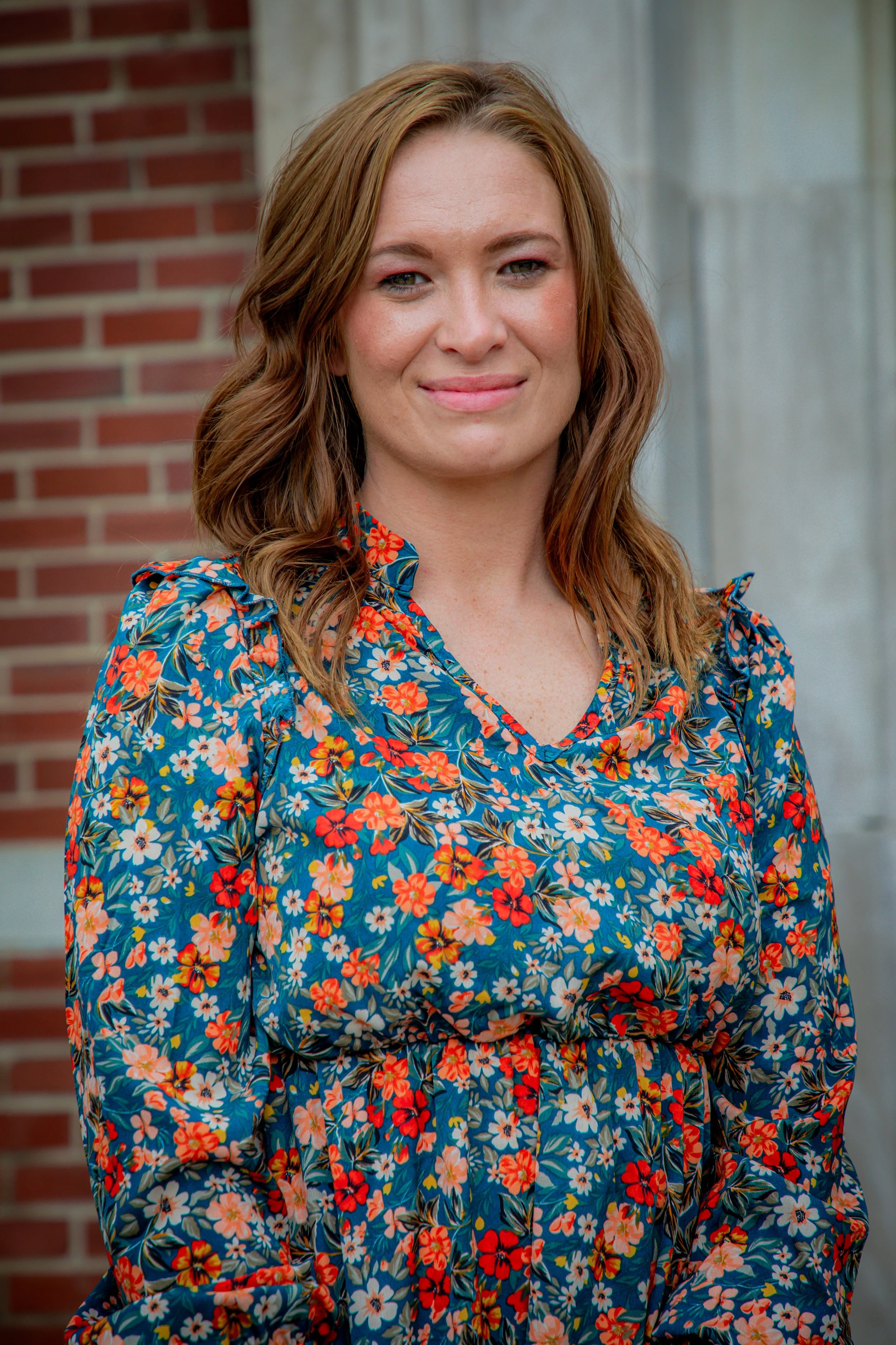 A woman with medium-length wavy red hair wearing a colorful floral dress, standing outdoors in front of a brick wall and a gray concrete structure.