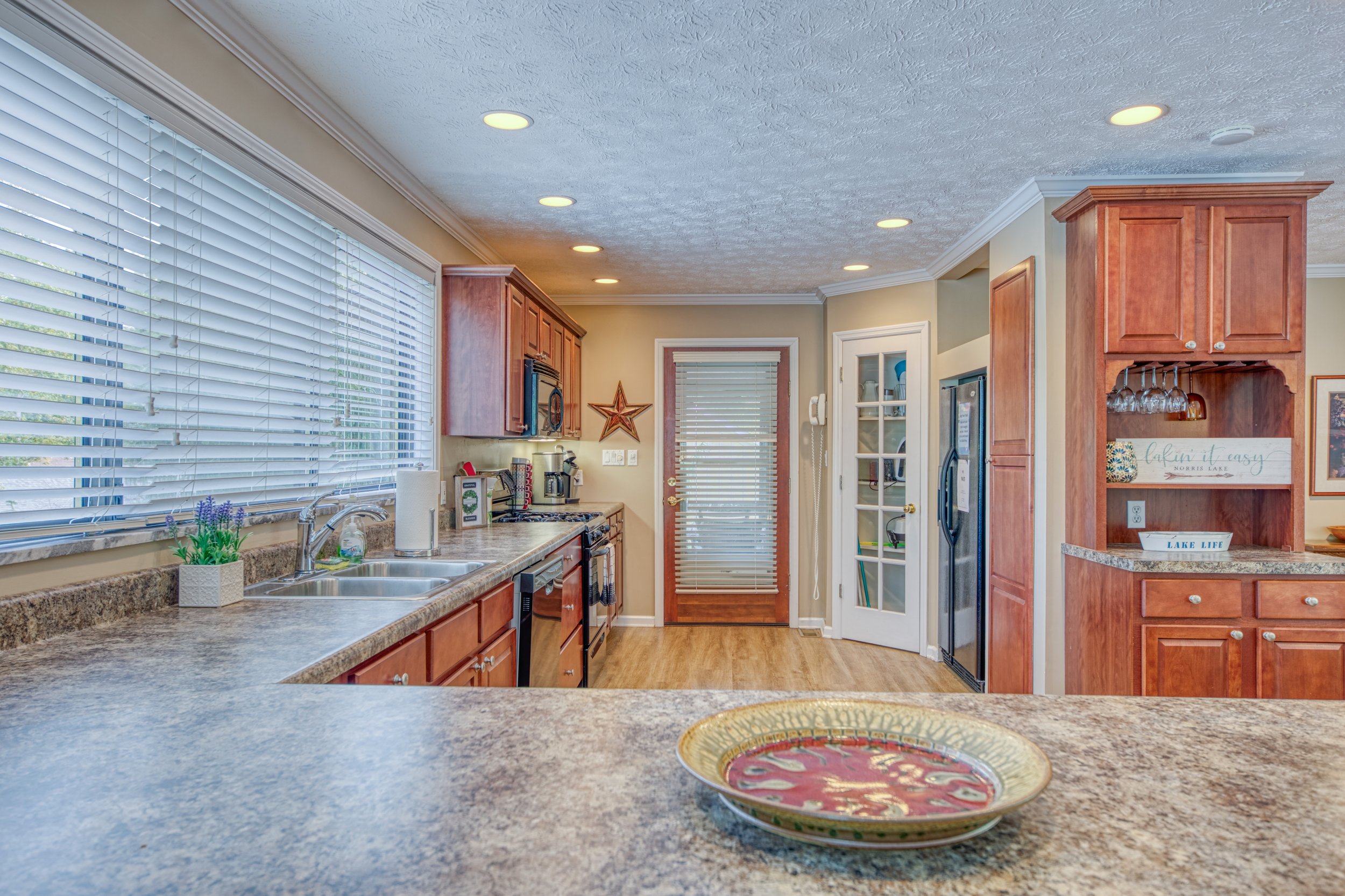 Kitchen with beige walls, wooden cabinets, granite countertops, window blinds, and decorative items including a star and a sign that says "Lakin it easy."
