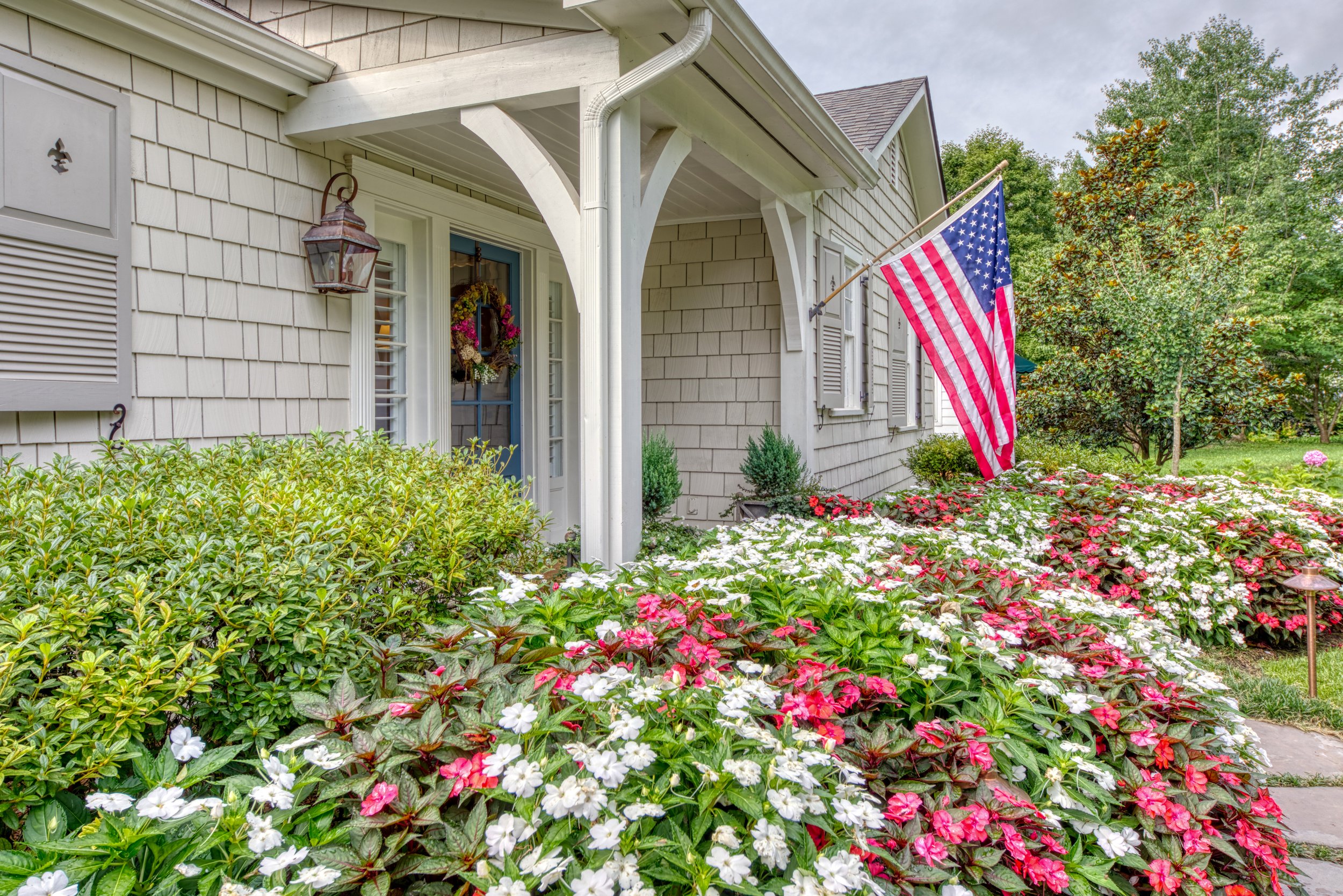 A front yard of a house with a garden filled with pink and white flowers, a bush, and a tree. The house has beige shingles, white trim, a blue front door with a floral wreath, a decorative lantern, and an American flag on a pole.