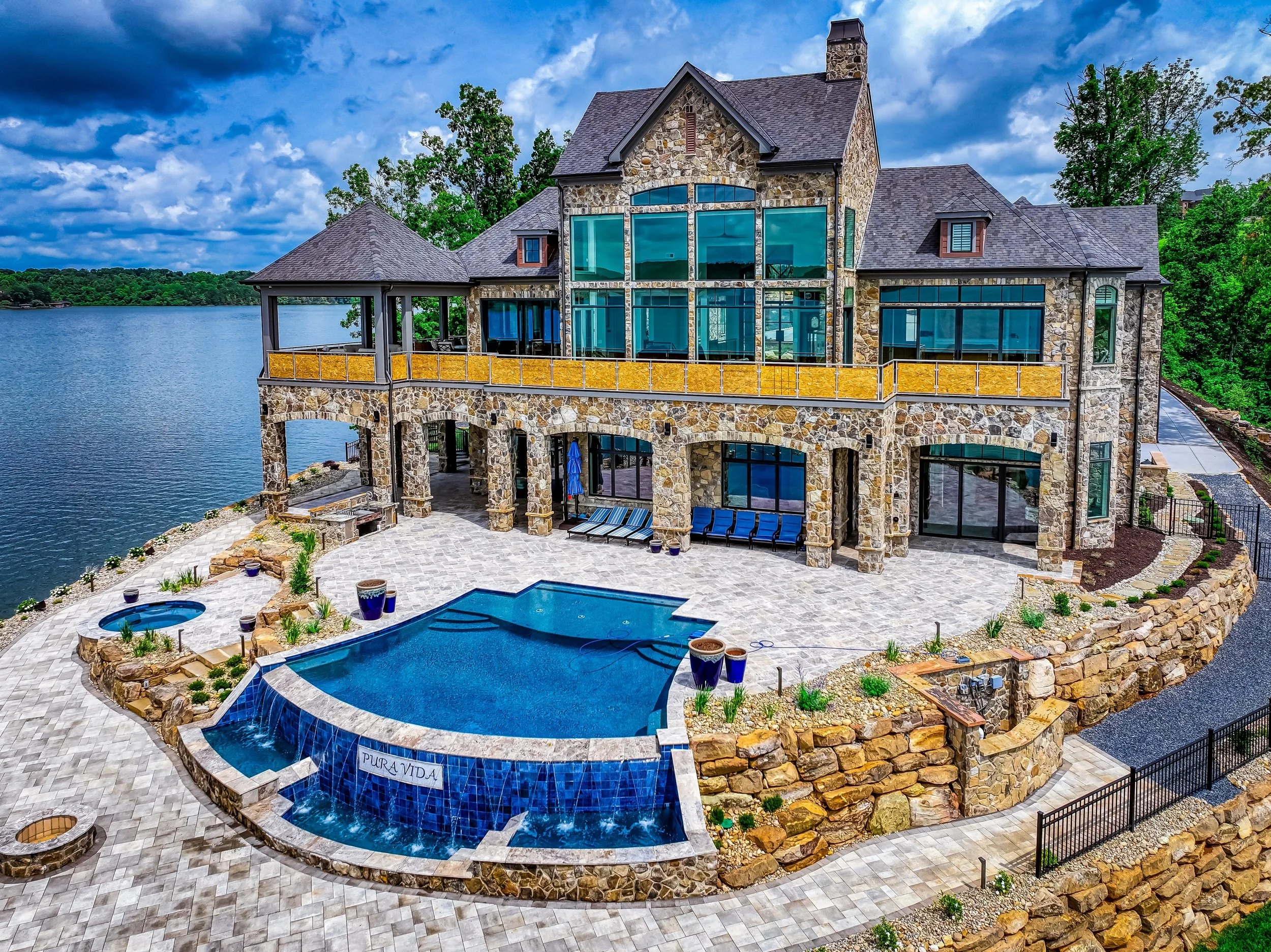 A large stone house with multiple rooflines, large blue-tinted windows, and a spacious balcony overlooking a lake. There is a backyard with a swimming pool and a hot tub, paved patio, and landscaped greenery.