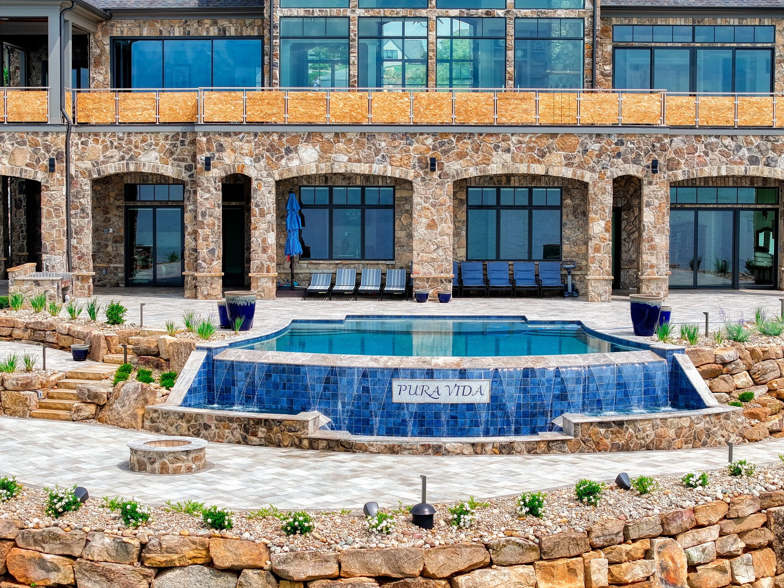 View of a multi-story stone and glass house with a pool in front and a stone-paved patio. The house has large windows and a balcony with an orange fence. There are chairs and an umbrella on the patio and potted plants around the pool. The name 'Pura 