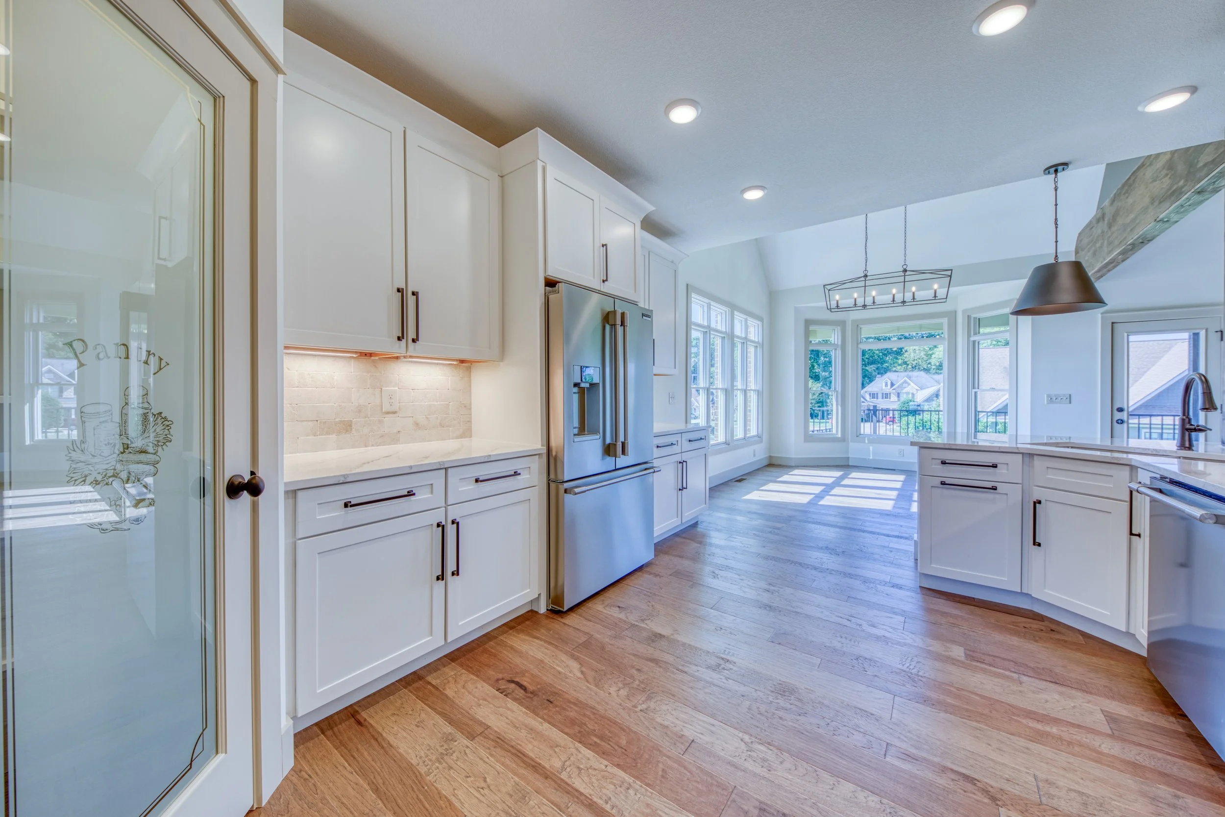 Bright modern kitchen with white cabinets, stainless steel refrigerator, large windows, wooden flooring, and contemporary pendant and chandelier lighting.