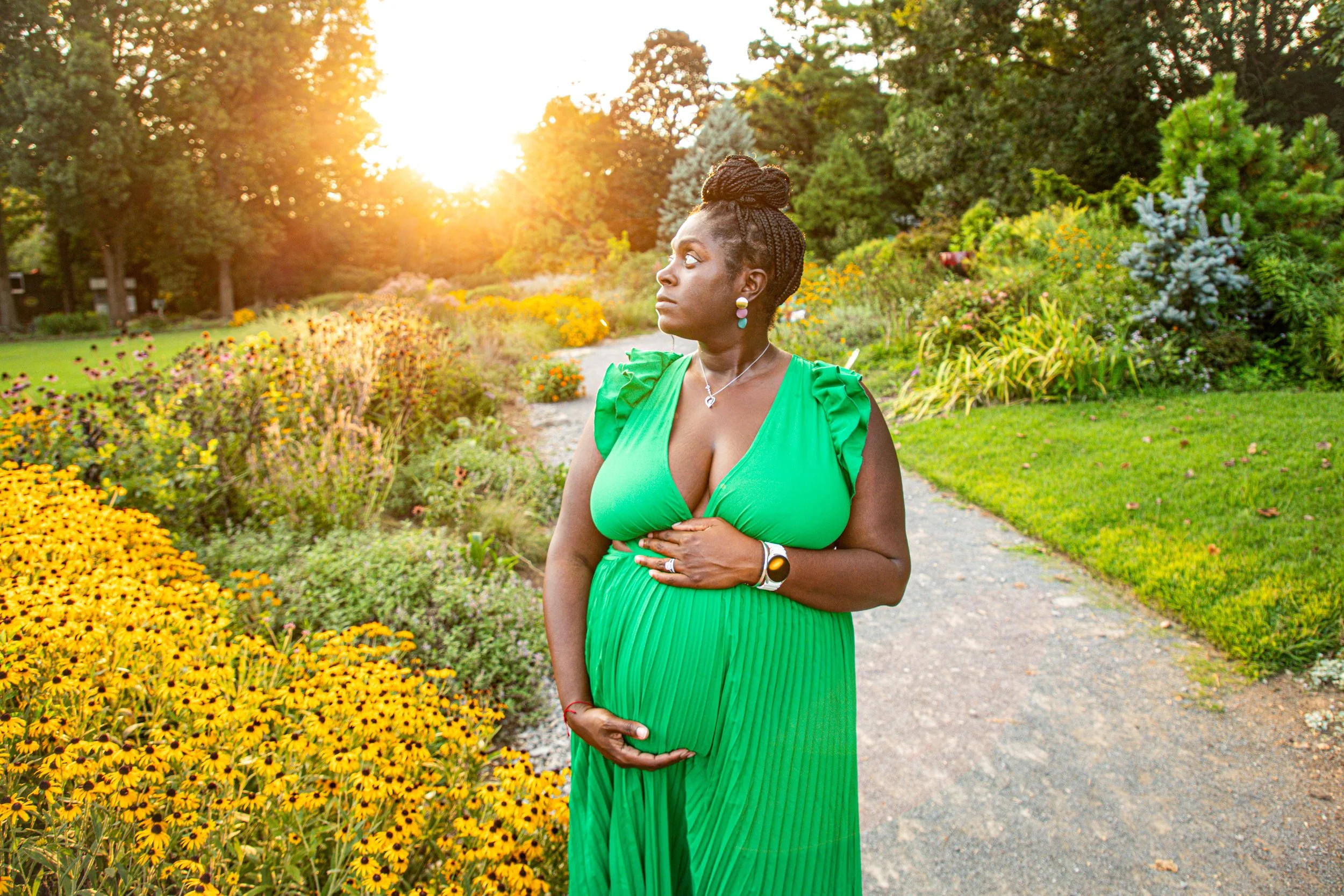 A pregnant woman in a green dress standing in a flower garden during sunset, with her hand resting on her belly and facing to the right.