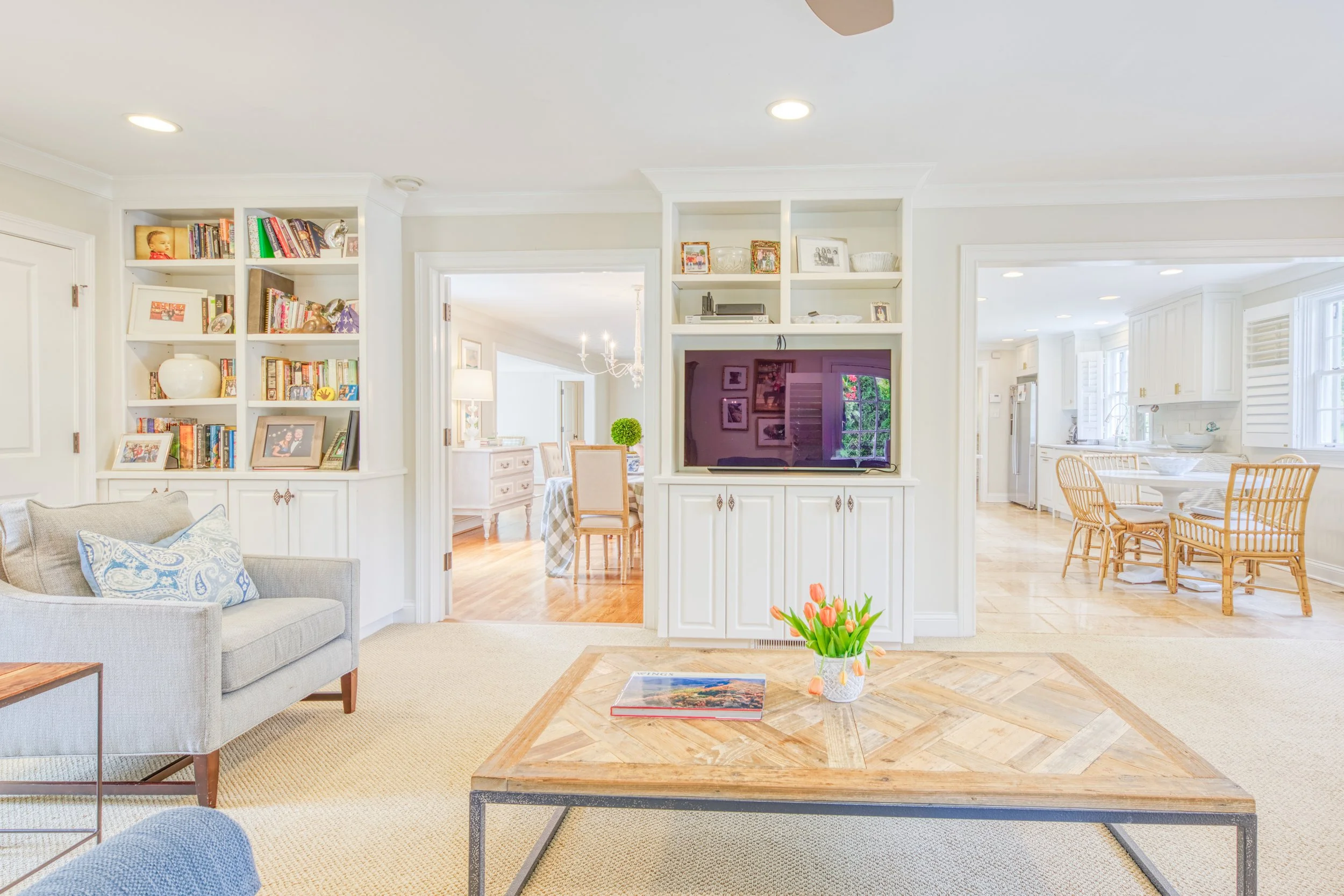 Living room with gray sofa, coffee table with book and vase of tulips, built-in bookshelf with books and photos, doorway to dining room with chandelier, open kitchen with white cabinets and dining table