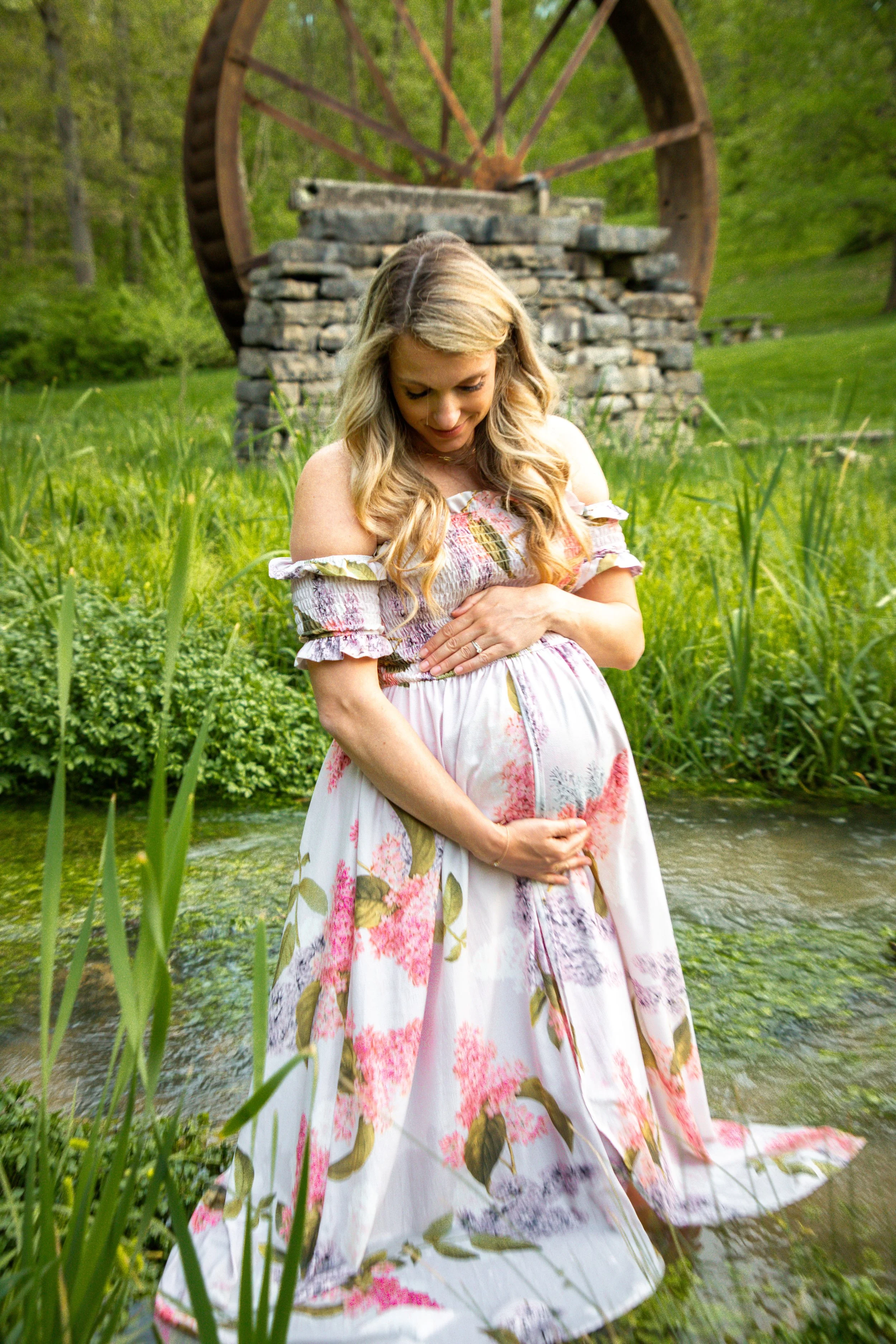 A pregnant woman standing in front of a stone watermill wheel in a lush green outdoor setting, gently holding her belly and smiling.
