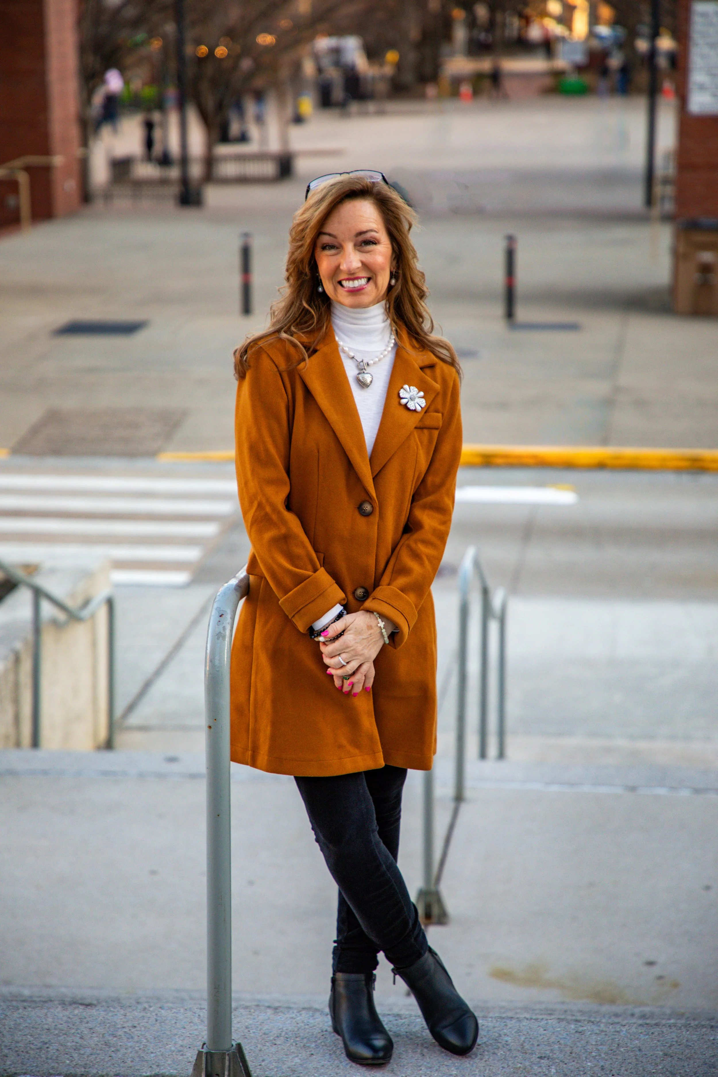 A smiling woman with light brown hair and pearl earrings, wearing a white turtleneck and mustard-colored coat, standing outdoors near a metal railing, with trees and buildings in the background during the evening.